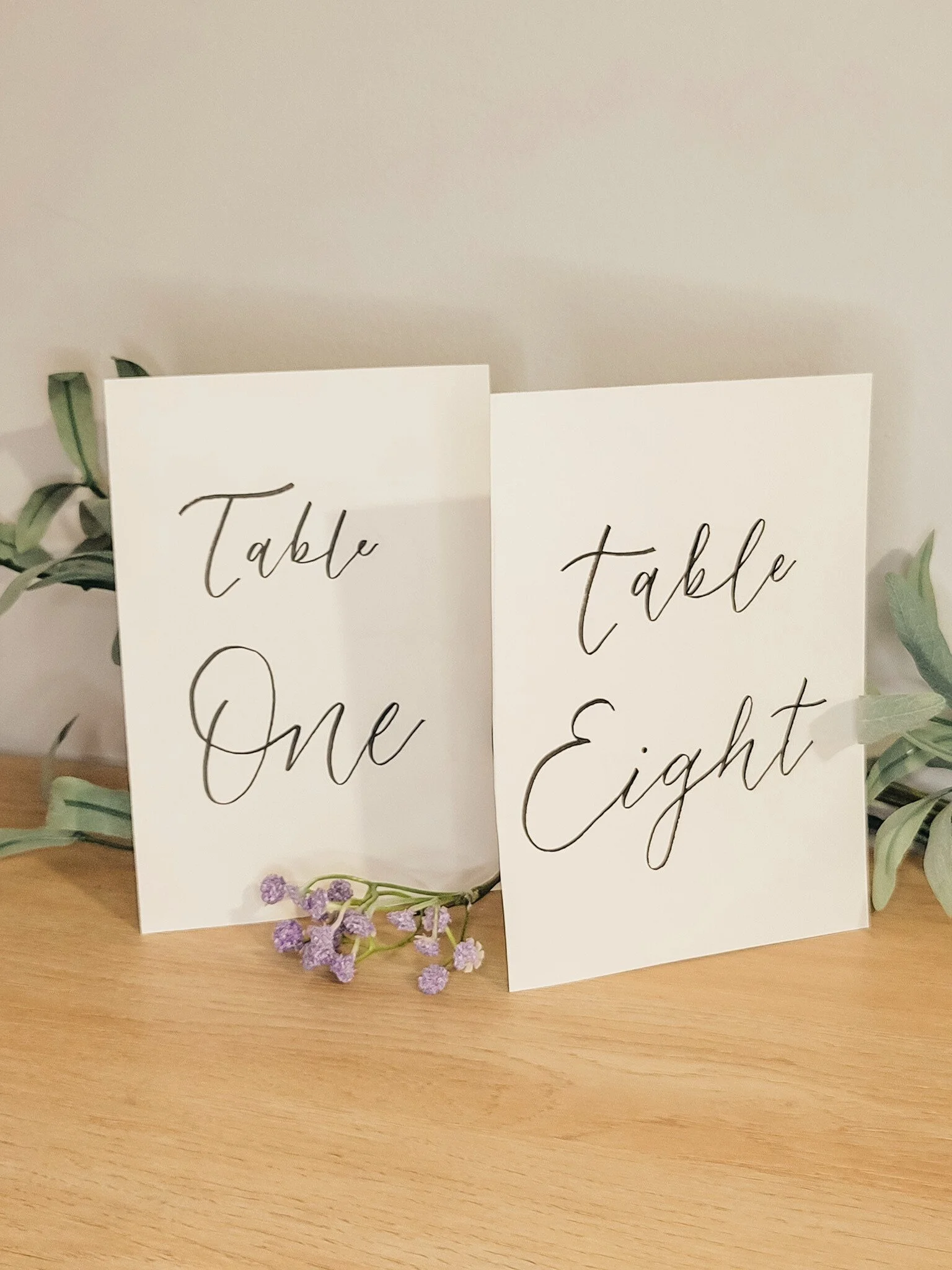 Hand-lettered wedding table numbers reading “Table One” and “Table Eight” on white card stock, styled with soft greenery and delicate lavender florals showcasing wedding stationery options.