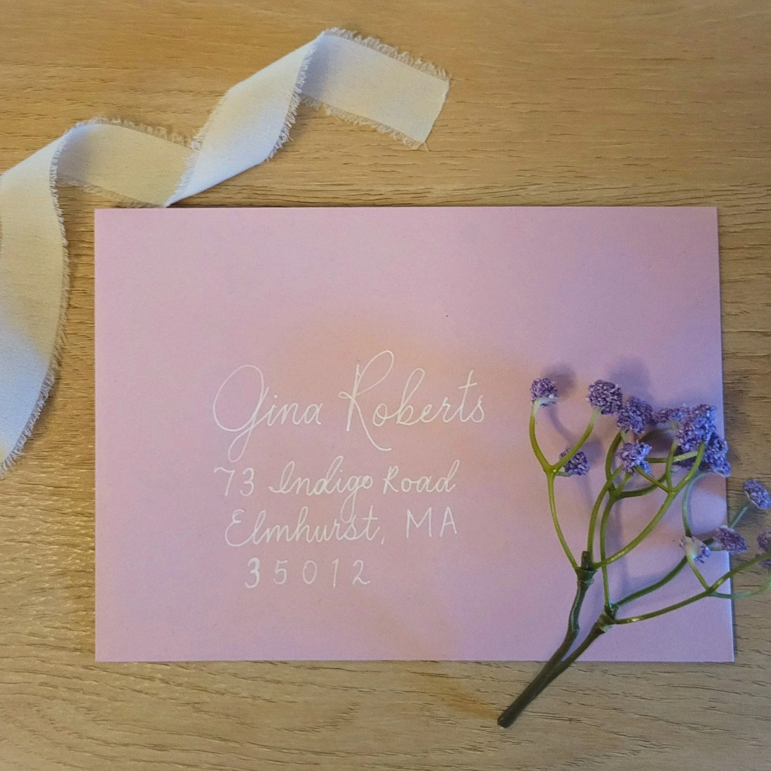 Custom envelope addressing in copperplate calligraphy. Pink envelope with white handwritten address, a white ribbon, and a small purple flower on a wooden surface.