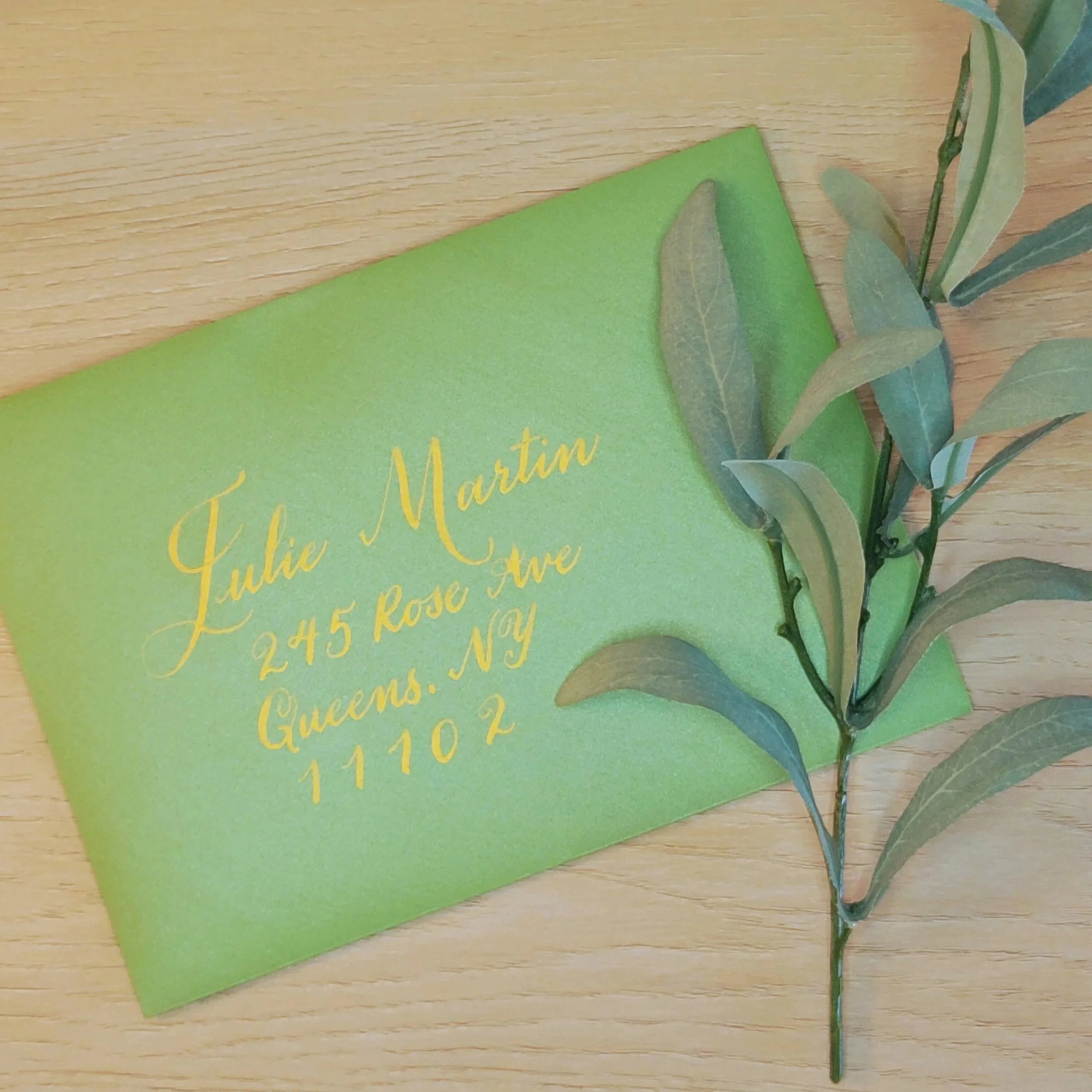 Personalized calligraphy envelope addressing displayed with a green envelope with gold ink and a branch with green leaves resting on a wooden surface.