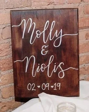 A custom calligraphy keepsake wooden sign with white hand-lettered couples' names 'Molly & Nicolas' which includes the wedding date '02-09-19' below. The sign is leaning against a brick wall with white cloth at the bottom.