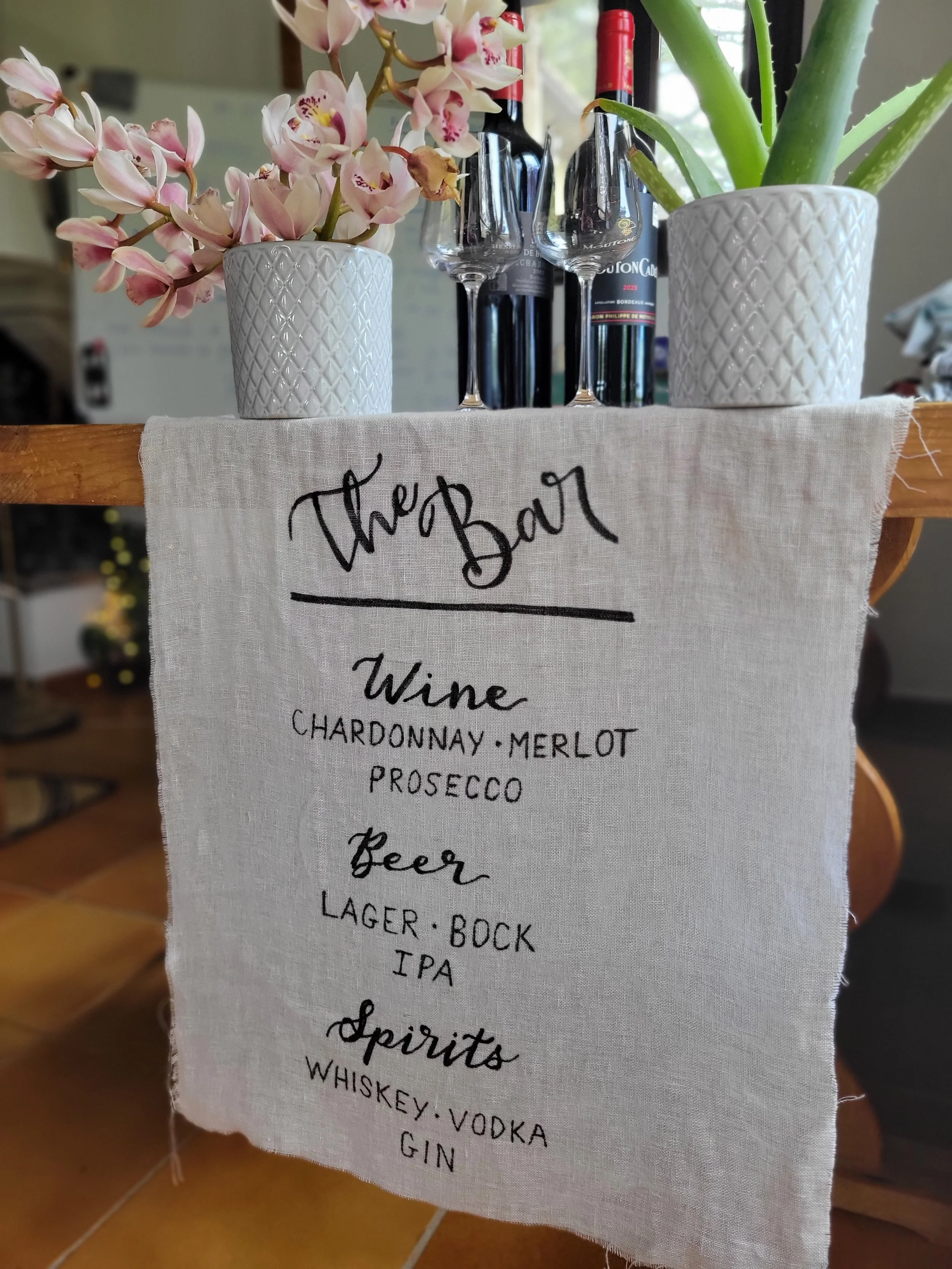 Hand-lettered linen bar menu sign reading "The Bar" with wine, beer, and spirits selections in calligraphy script, styled with orchids and wine bottles. Custom wedding and event signage by À La Court Calligraphy, Nice, Côte d'Azur.