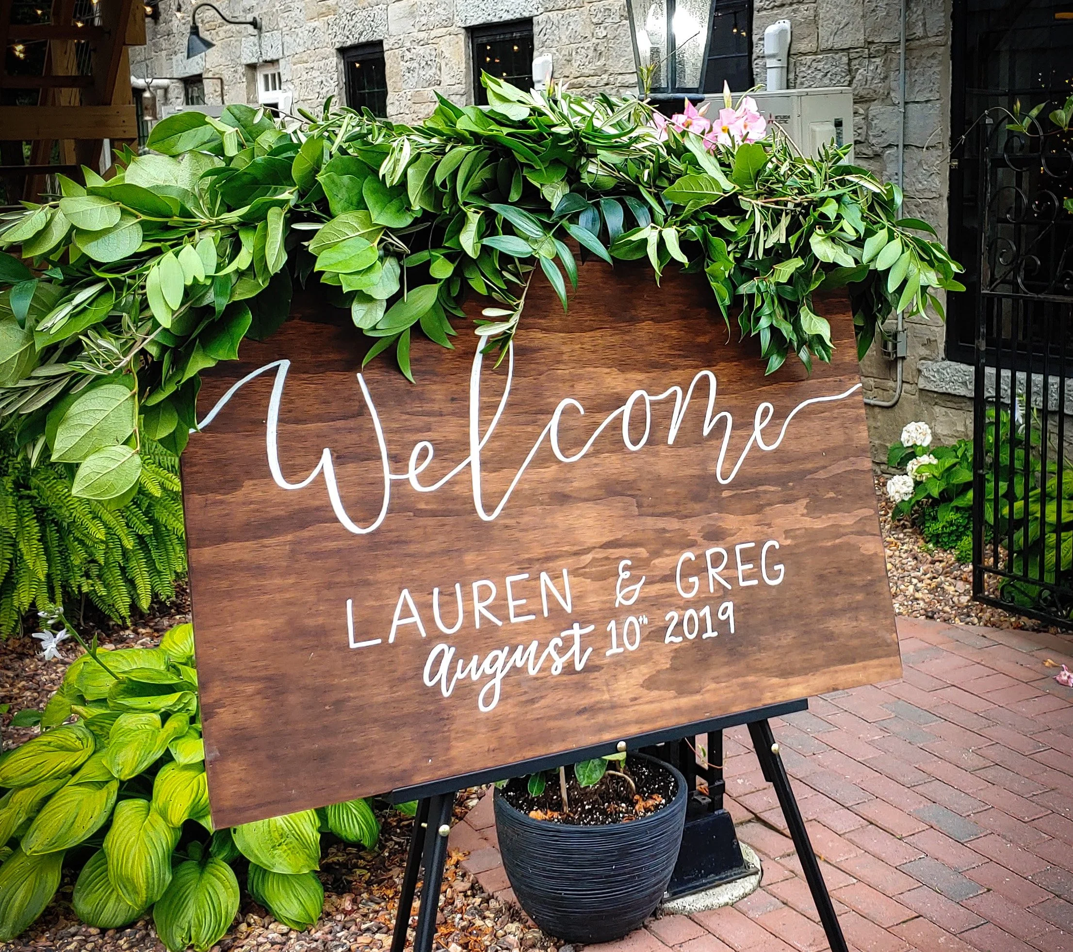 Custom wooden welcome sign with white hand-lettered text reading, 'Welcome Lauren & Greg August 10th 2019,' decorated with lush green leaves and placed outdoors on an easel with a brick pathway and a stone building in the background.