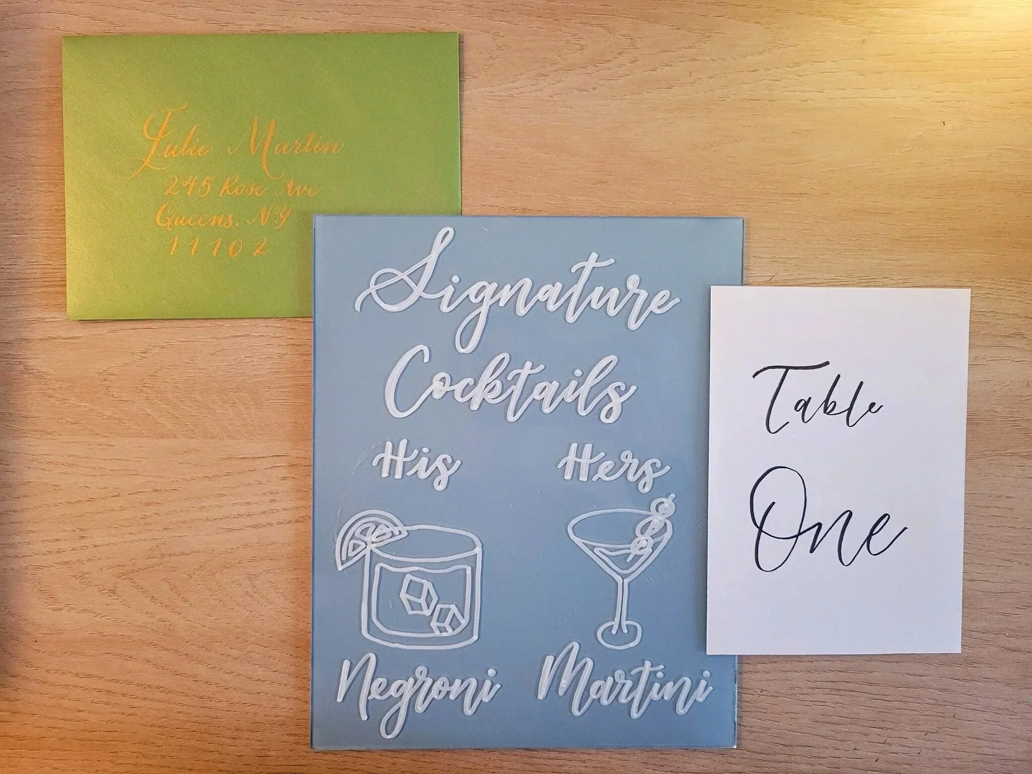 Three custom handwritten and handcrafted calligraphy pieces on a table: a green envelope addressed in gold ink ; a blue acrylic signature cocktails sign with drawings of a Martini and Negroni; and a table number sign in black in on ivory cardstock.