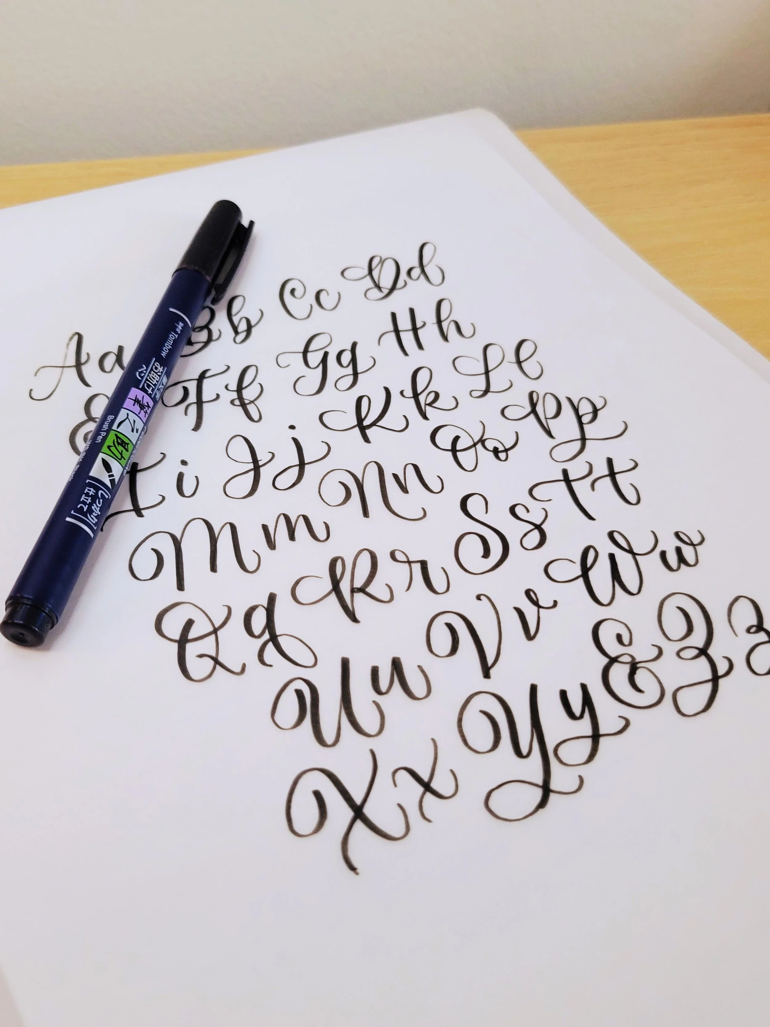 Sheet of paper with handwritten calligraphy alphabet in black ink, with a black calligraphy pen resting on top.