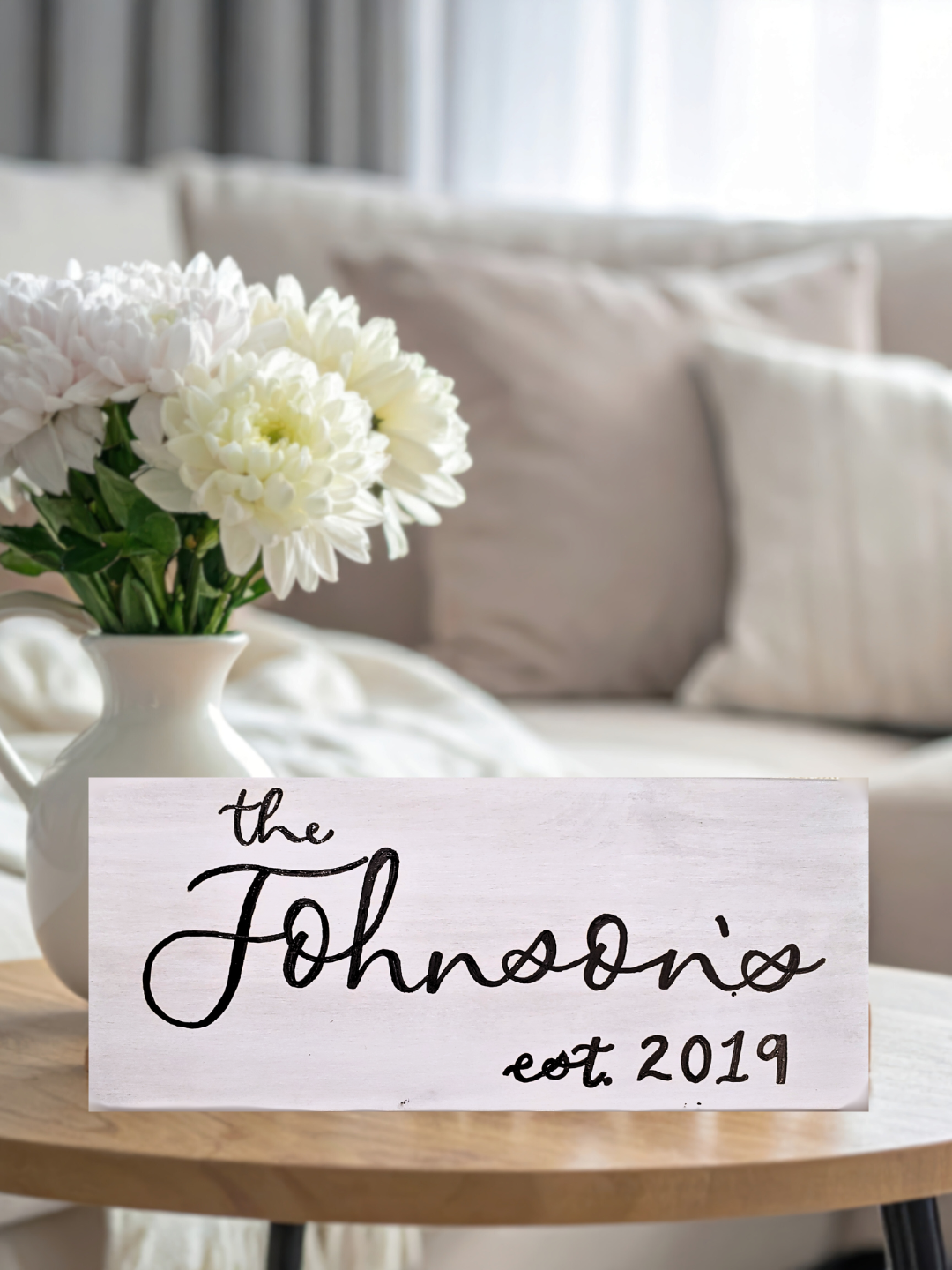 A custom calligraphy piece, white-washed wooden sign that reads "the Johnsons est. 2019" in black hand-lettering, placed on a light wooden table with a white vase of white flowers, and a beige sofa with light-colored pillows in the background.