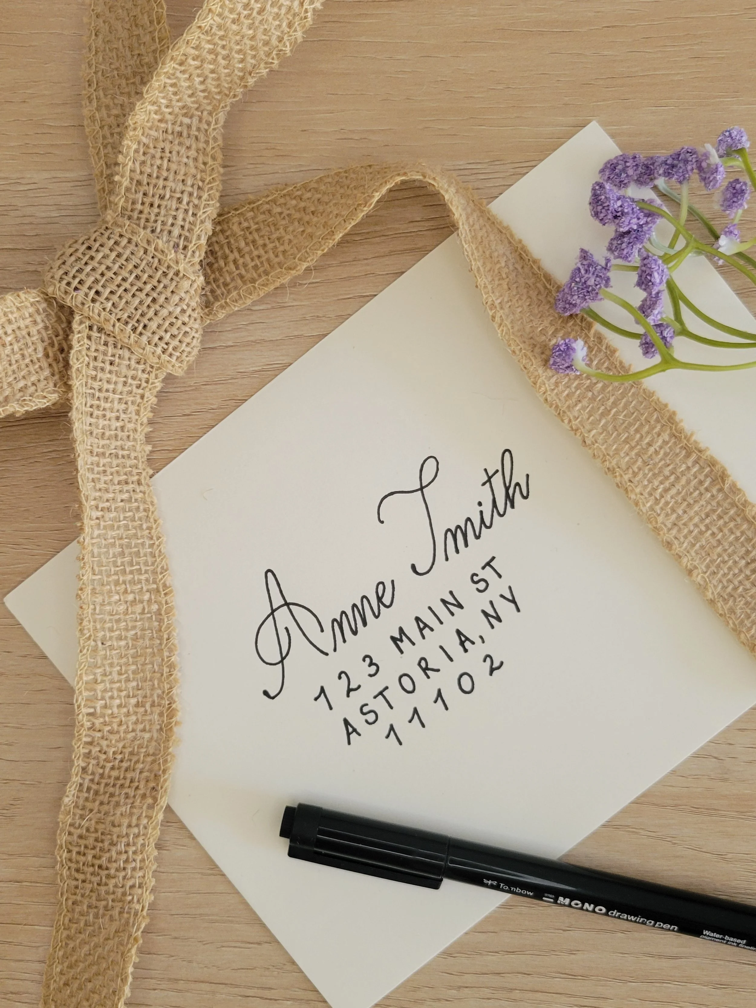 Custom calligraphy envelope with handwritten address: 'Anne Smith, 723 Main St, Astoria, NY 1102,' with a black pen placed on it, a decorative ribbon, and purple flowers on a wooden surface.