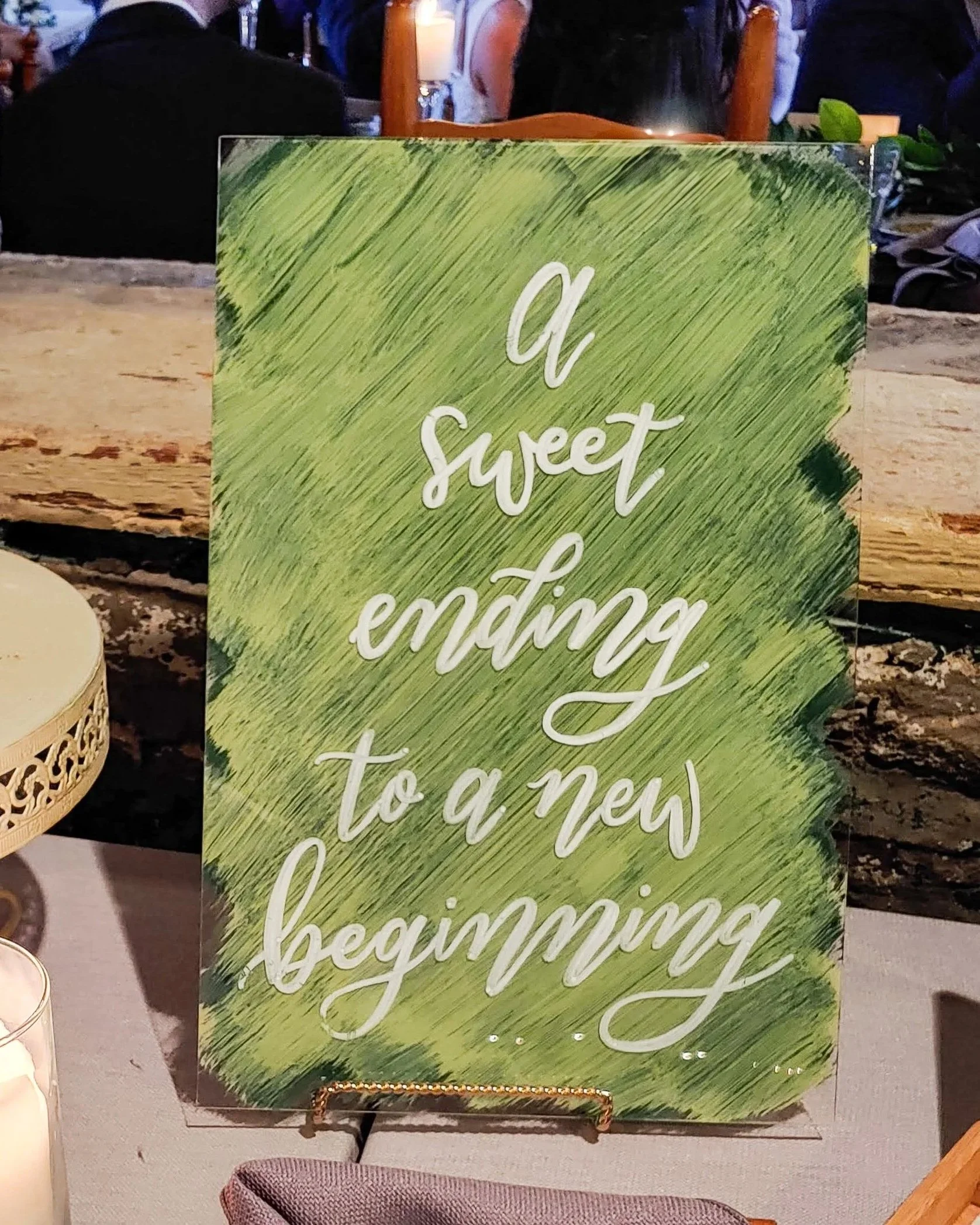 Decorative acrylic wedding dessert table sign with a green painted background and white calligraphy that reads 'A sweet ending to a new beginning'.