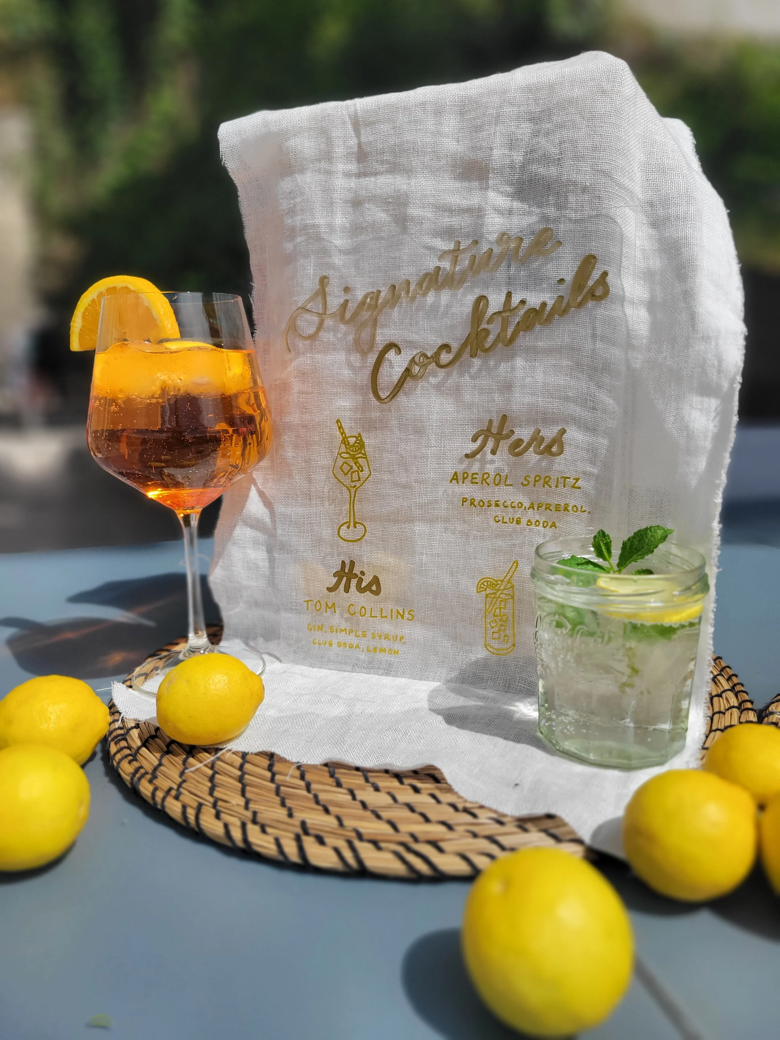 Hand-lettered linen signature cocktails sign in gold calligraphy script, styled with an Aperol Spritz, Tom Collins, and fresh lemons on a wicker tray outdoors. Custom wedding bar signage by À La Court Calligraphy, Nice, French Riviera.