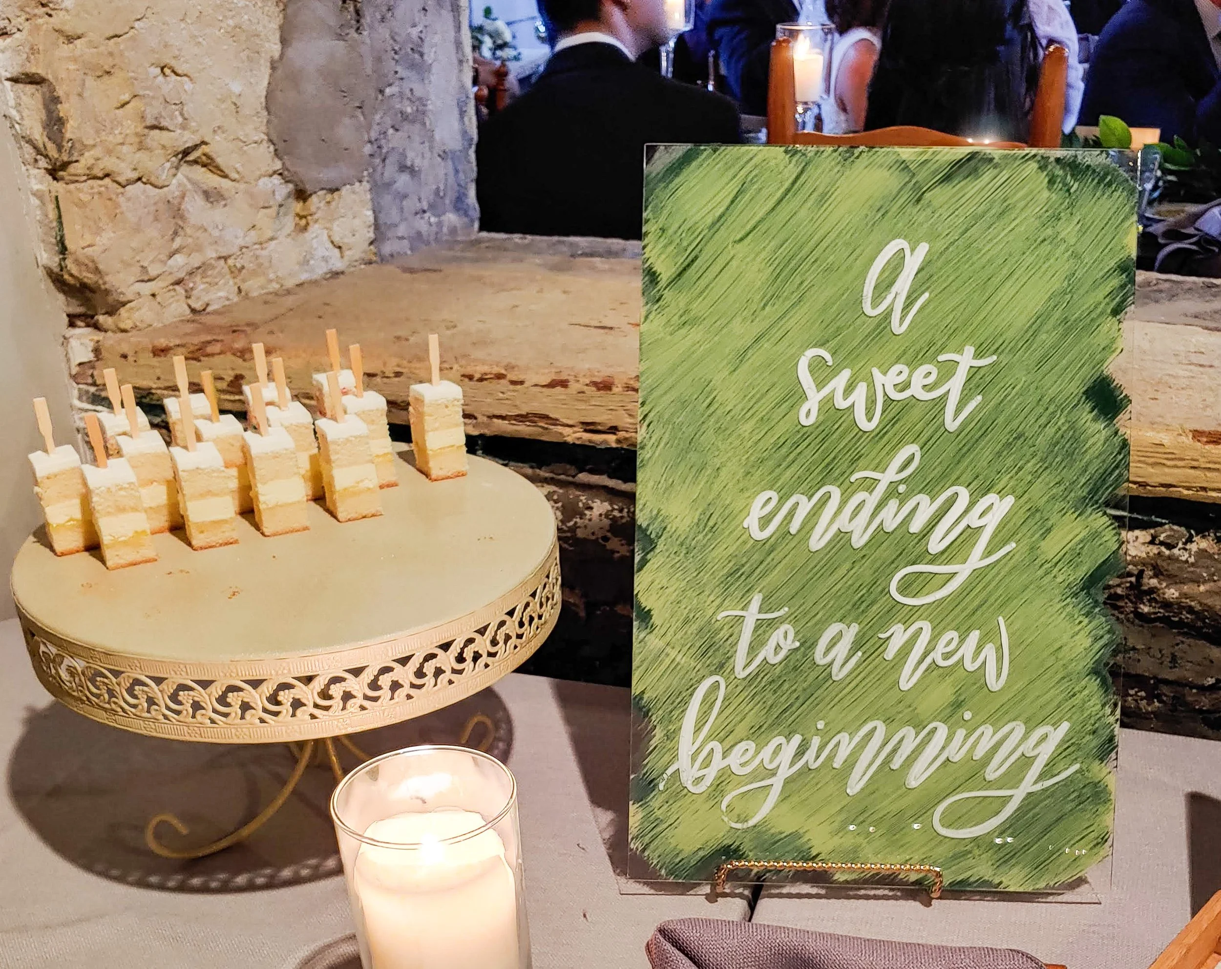 Small cake pops with white and yellow layers on a gold decorative stand, with a candle in front. 	
Decorative acrylic wedding dessert table sign with a green painted background and white calligraphy that reads 'A sweet ending to a new beginning'.