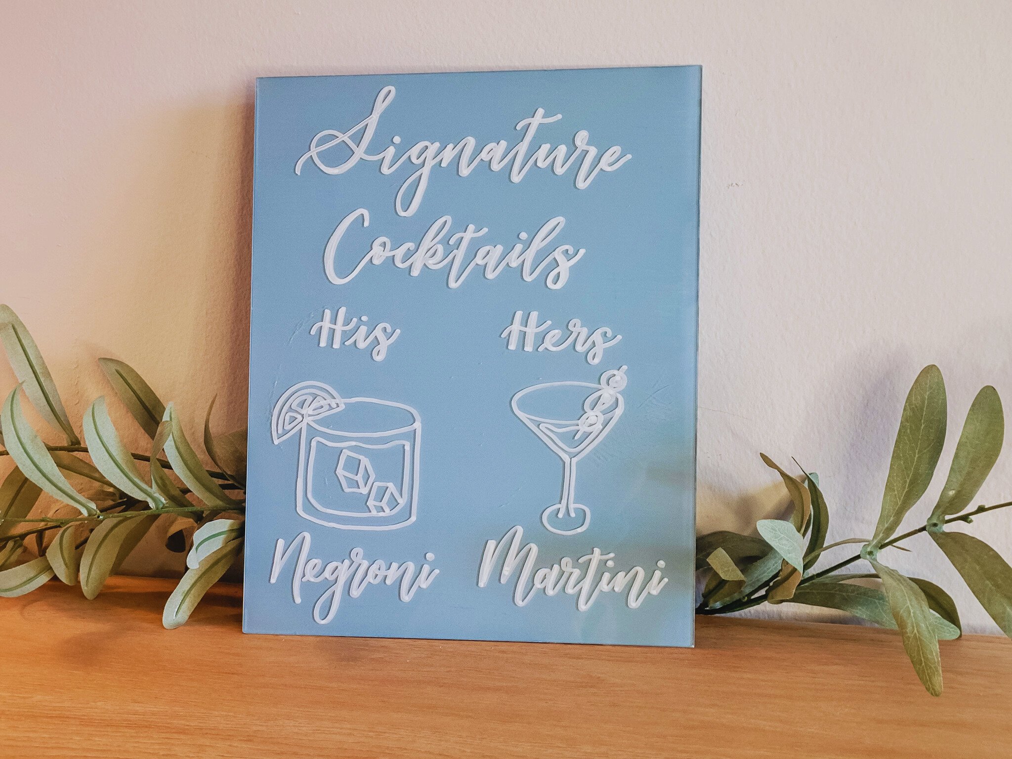 Bespoke acrylic signature cocktails bar sign with hand-lettered text and hand-drawn illustrations of a Negroni and a Martini, placed on a wooden surface with green foliage.