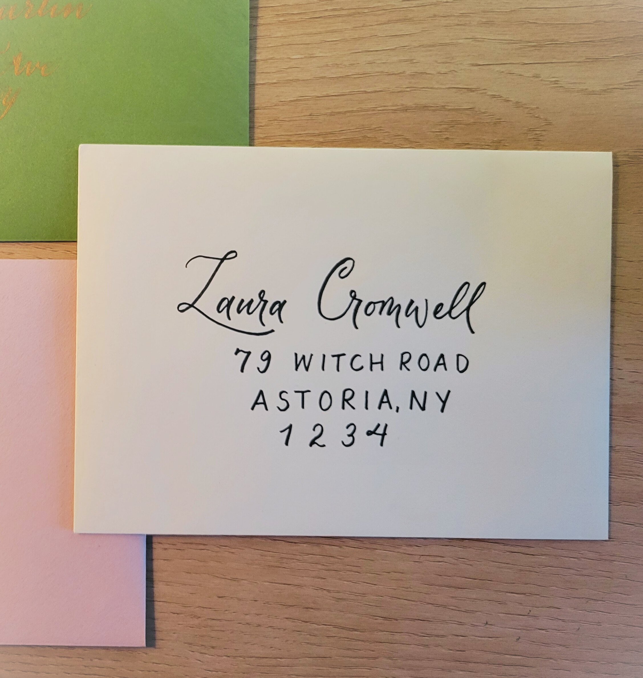 A white custom envelope with handwritten calligraphy in black ink displaying an address: 'Laura Cromwell, 79 Witch Road, Astoria, NY, 1234'. The card is on a wooden surface, with parts of other cards in pink and green visible in the corners.