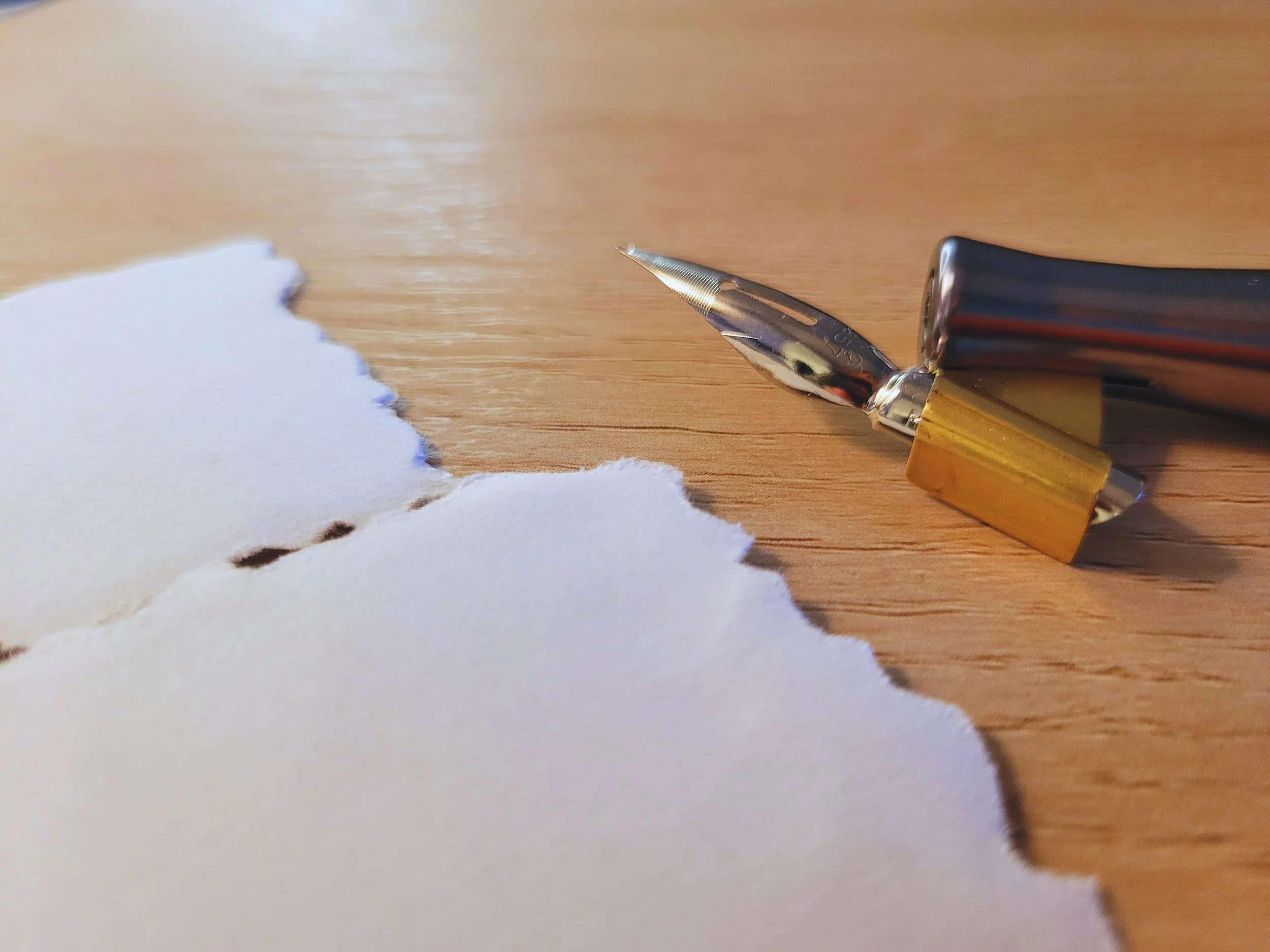 Close-up of a dip pen with a gold and silver body, resting on a wooden surface next to deckle edge ivory cardstock.