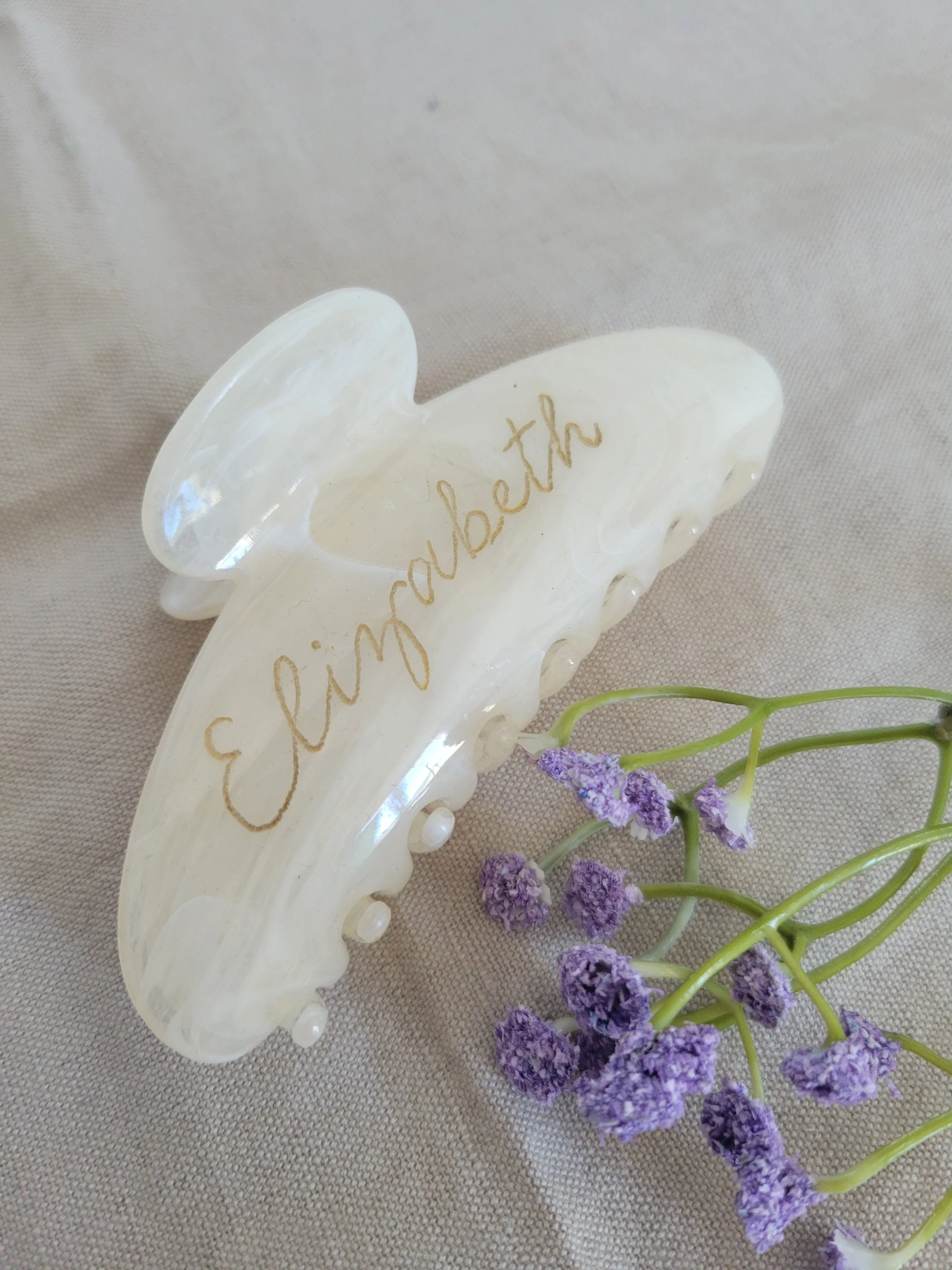 Hand-engraved pearl white claw hair clip with the name "Elizabeth" in gold calligraphy script, styled with dried purple flowers on a linen background. Personalized bridesmaid gift by À La Court Calligraphy, French Riviera.