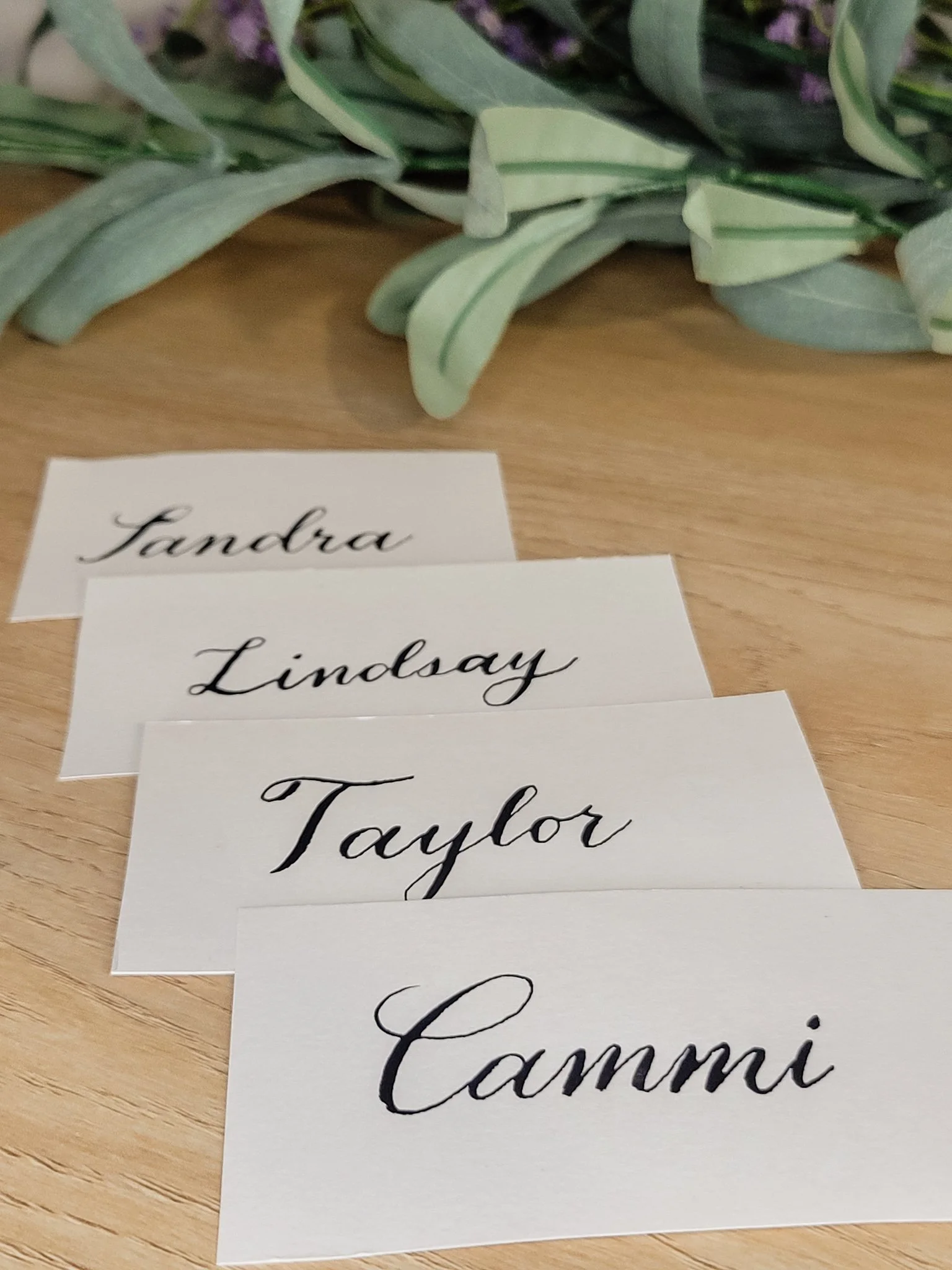 Custom hand-lettered wedding place cards with guest names written in black ink calligraphy, arranged on a wooden table with soft greenery accents.