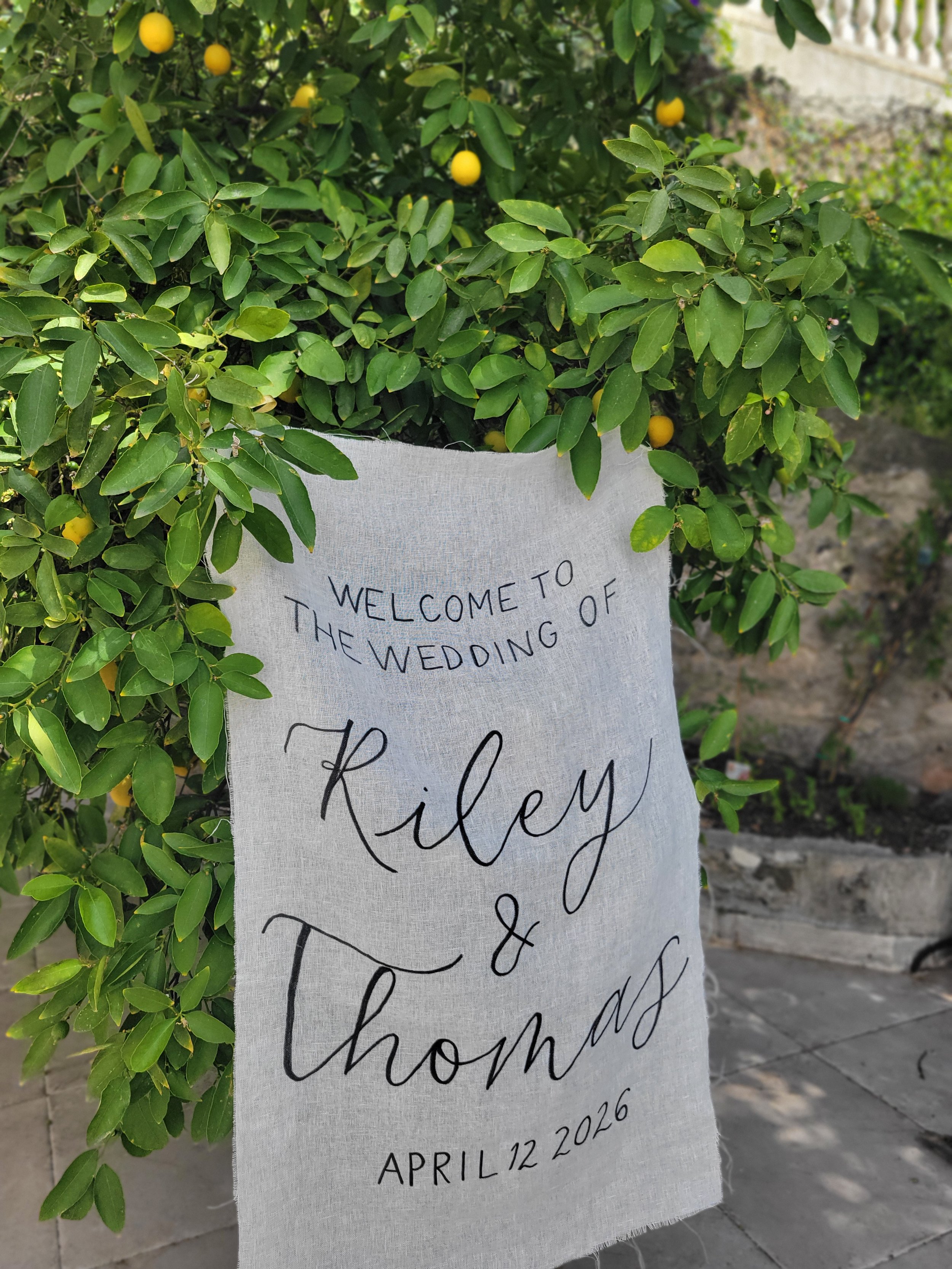 Hand-lettered linen wedding welcome sign reading "Welcome to the Wedding of Riley & Thomas, April 12 2026", hung outdoors against a lemon tree. Custom fabric wedding signage by À La Court Calligraphy, French Riviera.