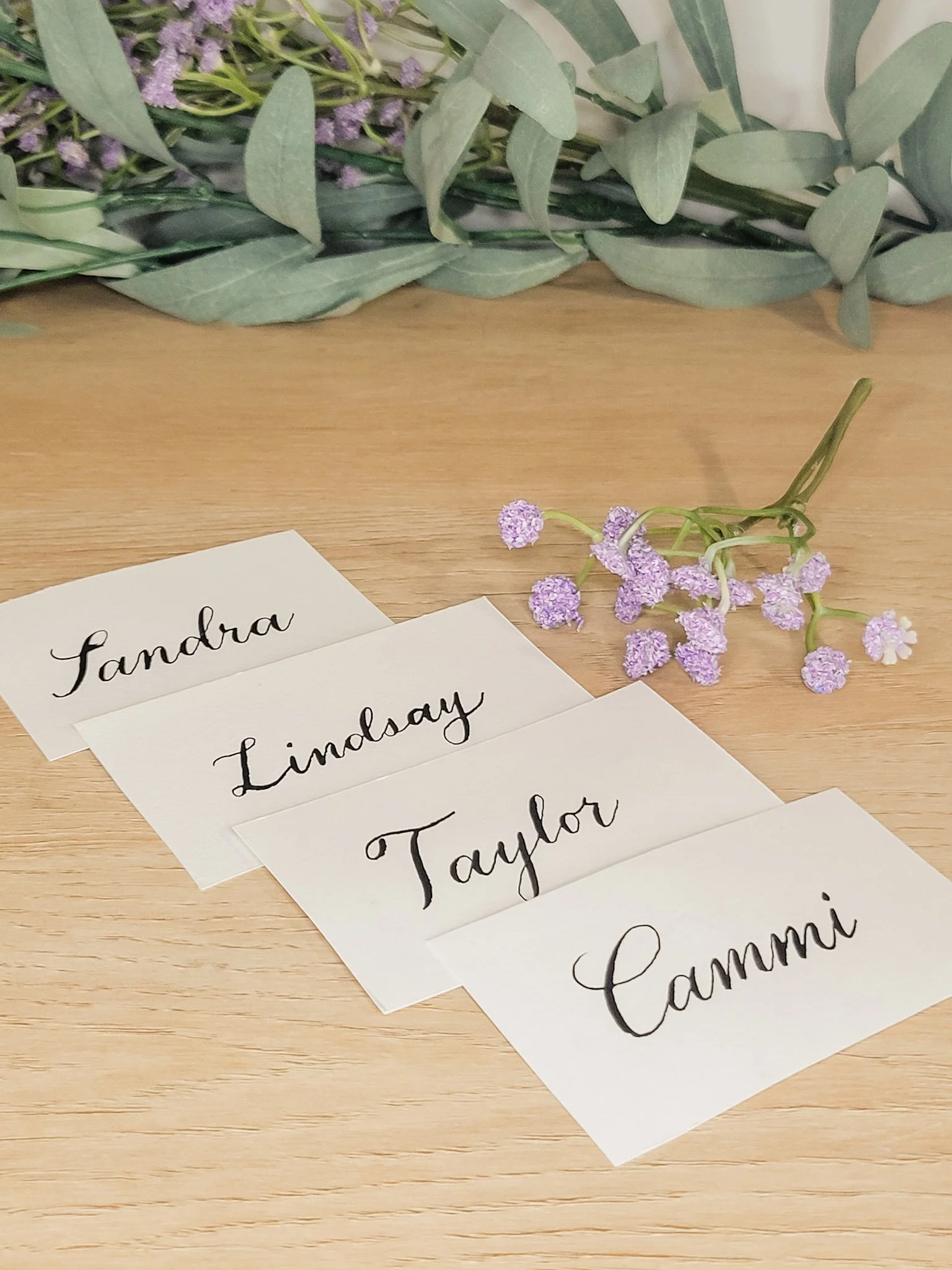 Four white wedding place cards with names handwritten in calligraphy with black ink: Sandra, Lindsay, Taylor, Cammi. A small sprig of purple flowers lies next to the papers on a wooden surface, with a backdrop of green leaves.