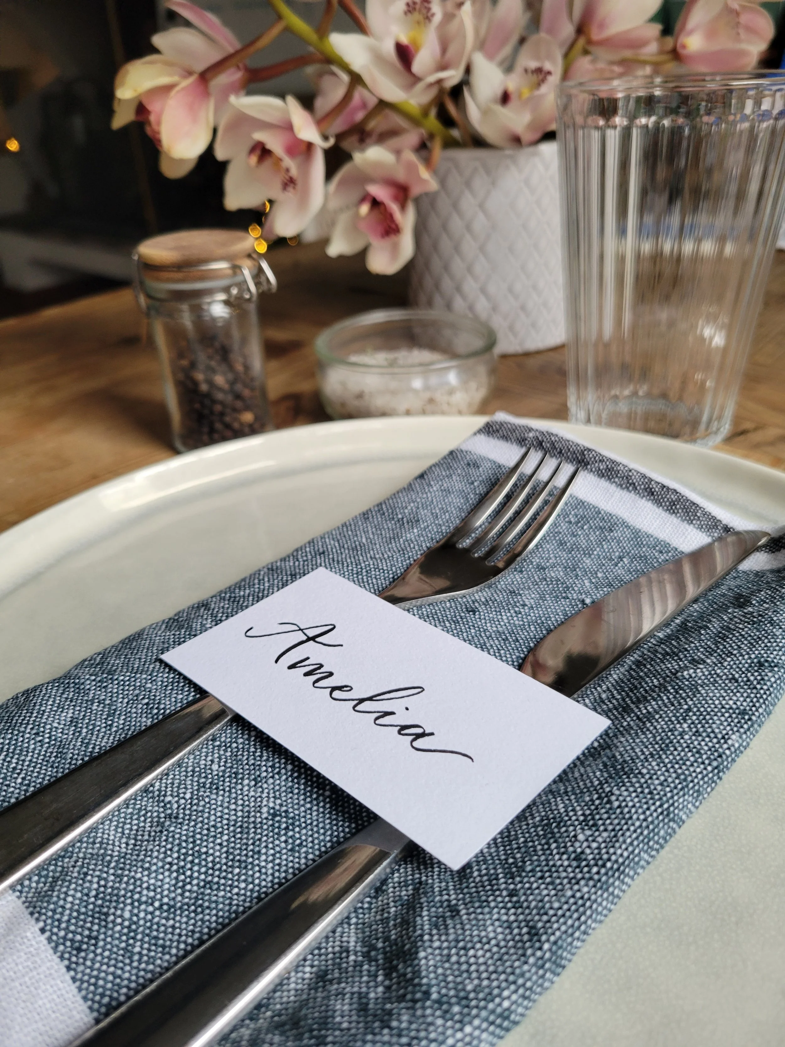 Hand-lettered calligraphy place card reading "Amelia" in black script, styled on a table setting with a linen napkin, silverware, and pink orchids. Custom wedding and event place cards by À La Court Calligraphy, Nice, French Riviera.