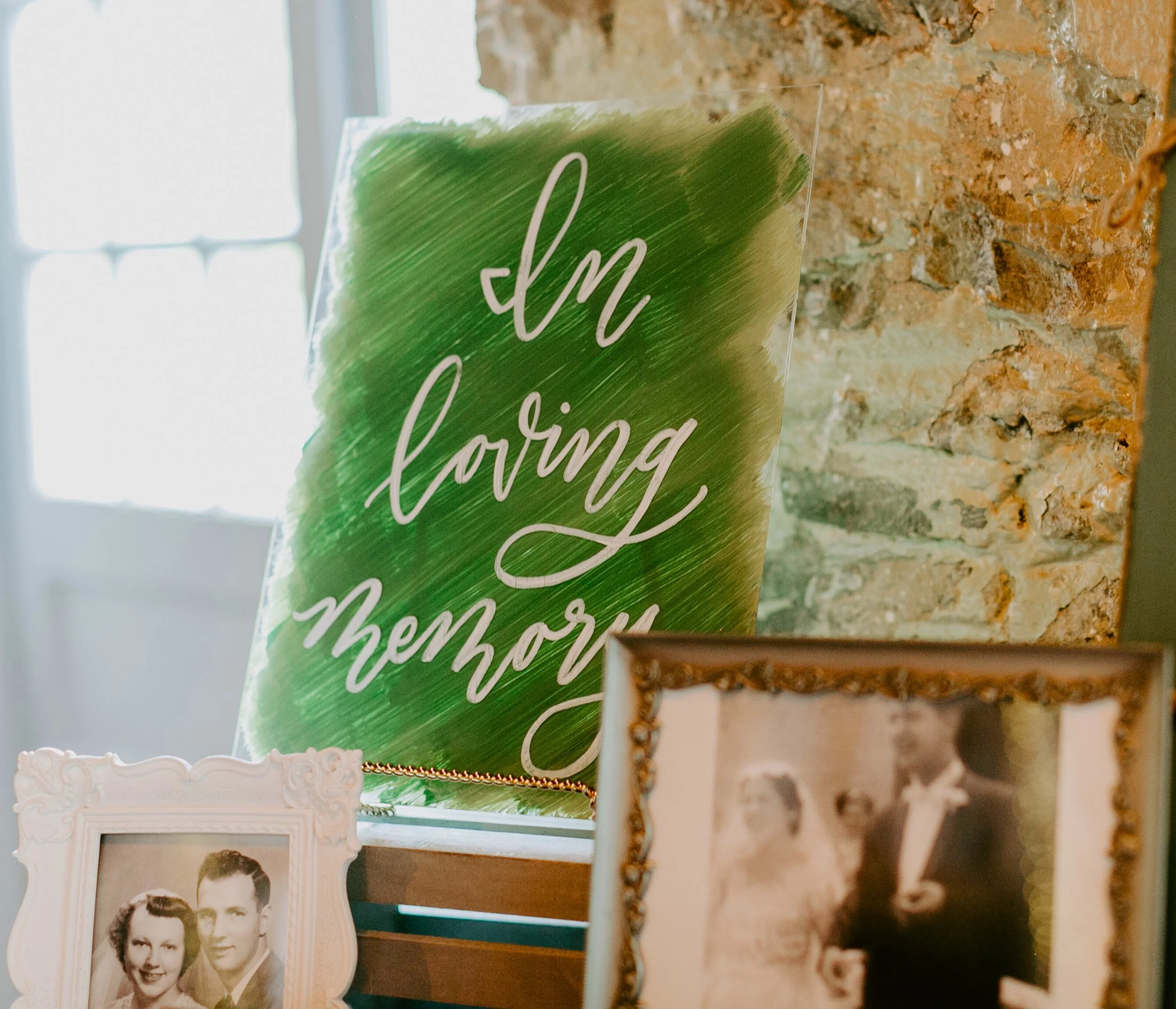 Clear acrylic remembrance sign with green painted background and hand-lettered text. Framed black-and-white photographs of a couple are in the foreground. The sign is placed in a rustic venue interior with brick wall.