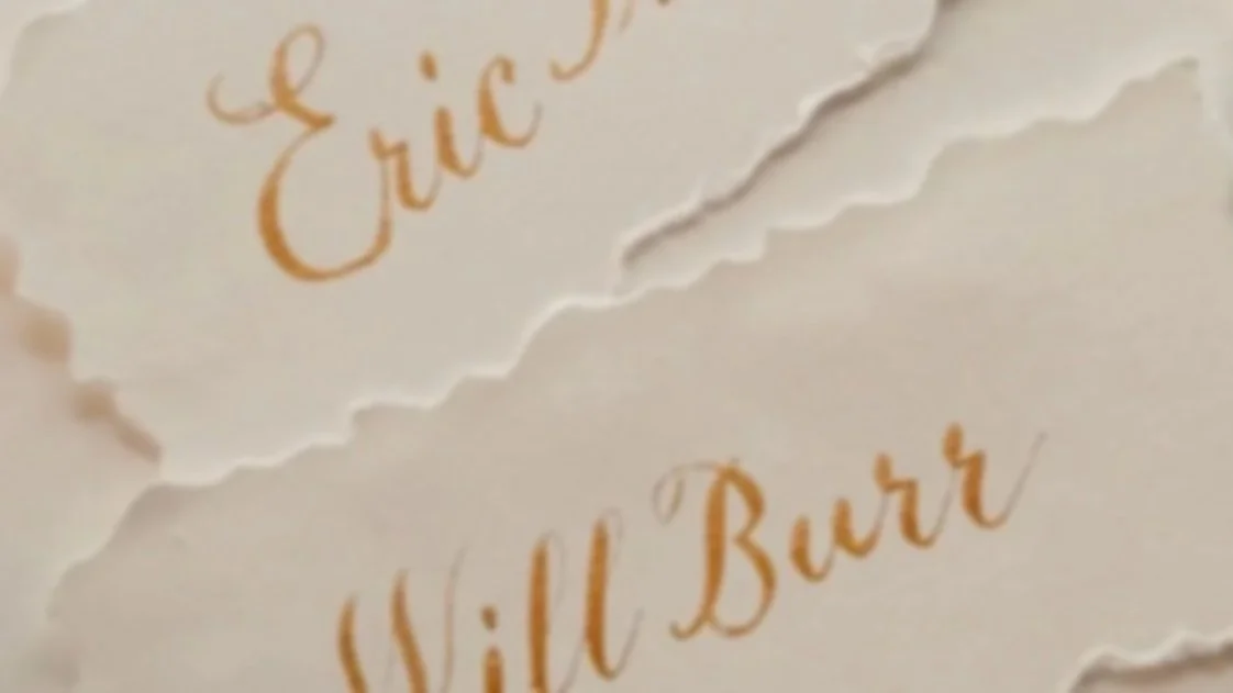 Close-up of deckle edge paper place cards with the names 'Eric' and 'Will Burr' written in gold ink copperplate calligraphy.