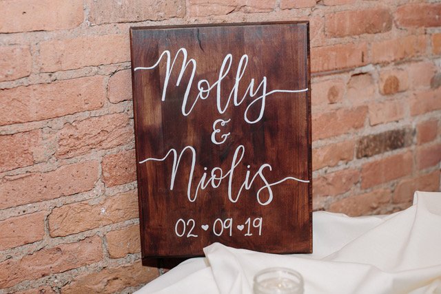 A custom calligraphy keepsake wooden sign with white hand-lettered couples' names 'Molly & Nicolas' which includes the wedding date '02-09-19' below. The sign is leaning against a brick wall with white cloth at the bottom.