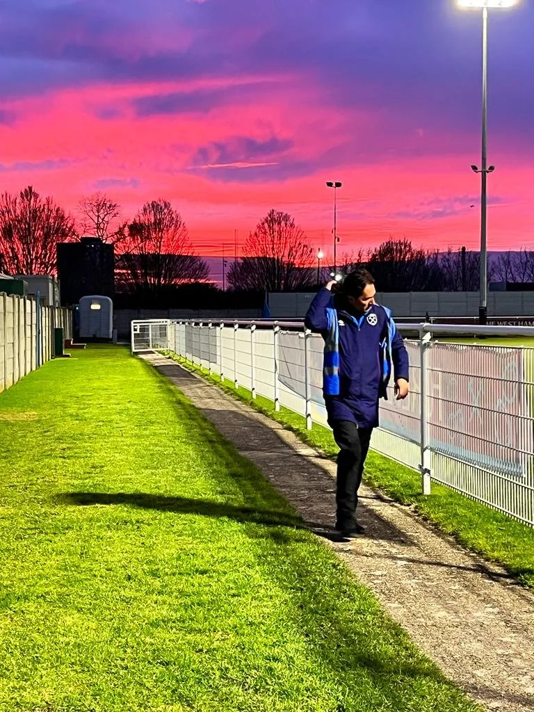 A man walking along a pathway beside a sports field with a pink and purple sunset sky in the background.