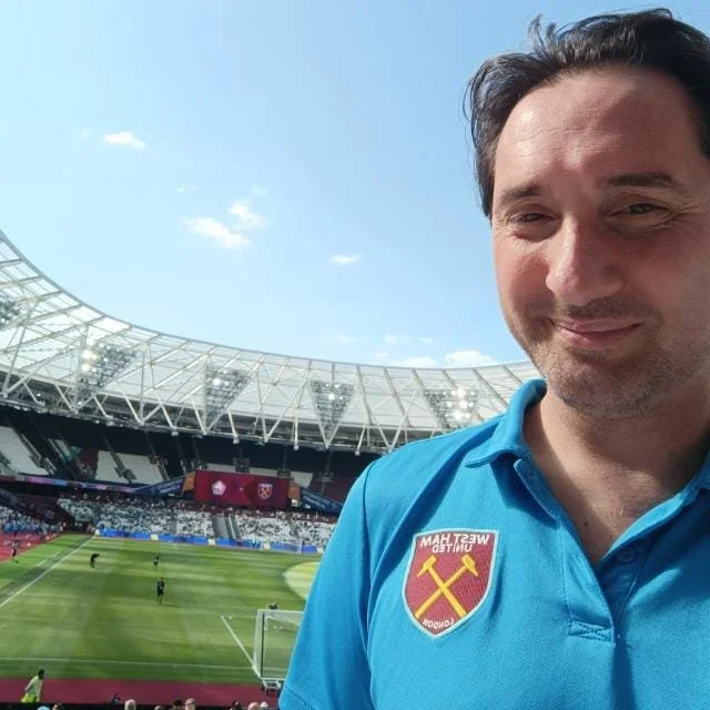 A man smiling at the camera in a stadium, wearing a West Ham United football shirt with the team's crest visible.