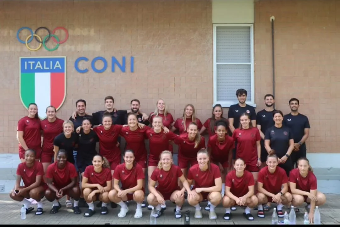 Group of female and male athletes in red and dark blue sportswear, posing outdoors in front of a wall with an Italian shield and Olympic rings, with the words 'ITALIA' and 'CONI' on the wall.