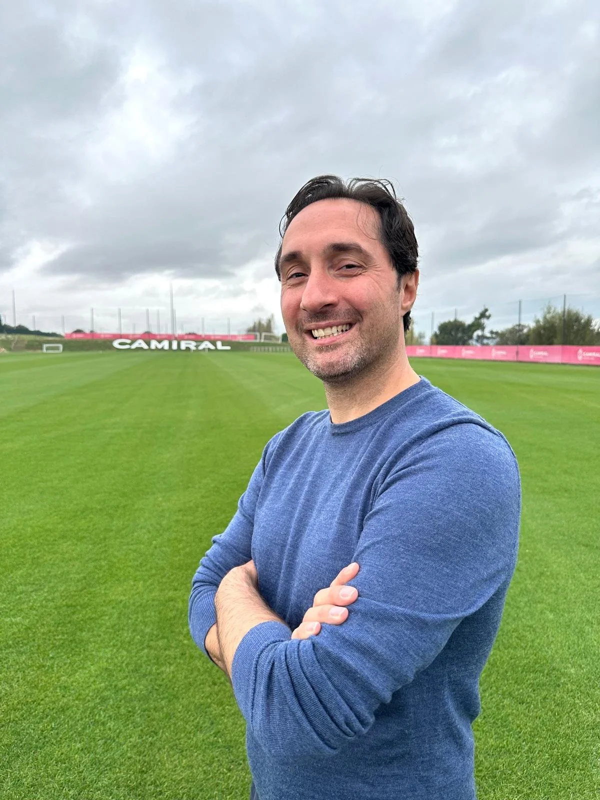 A man in a blue shirt standing with arms crossed on a grassy sports field with cloudy sky, pink advertising boards, and a sign that says 'CAMIRAL' in the background.