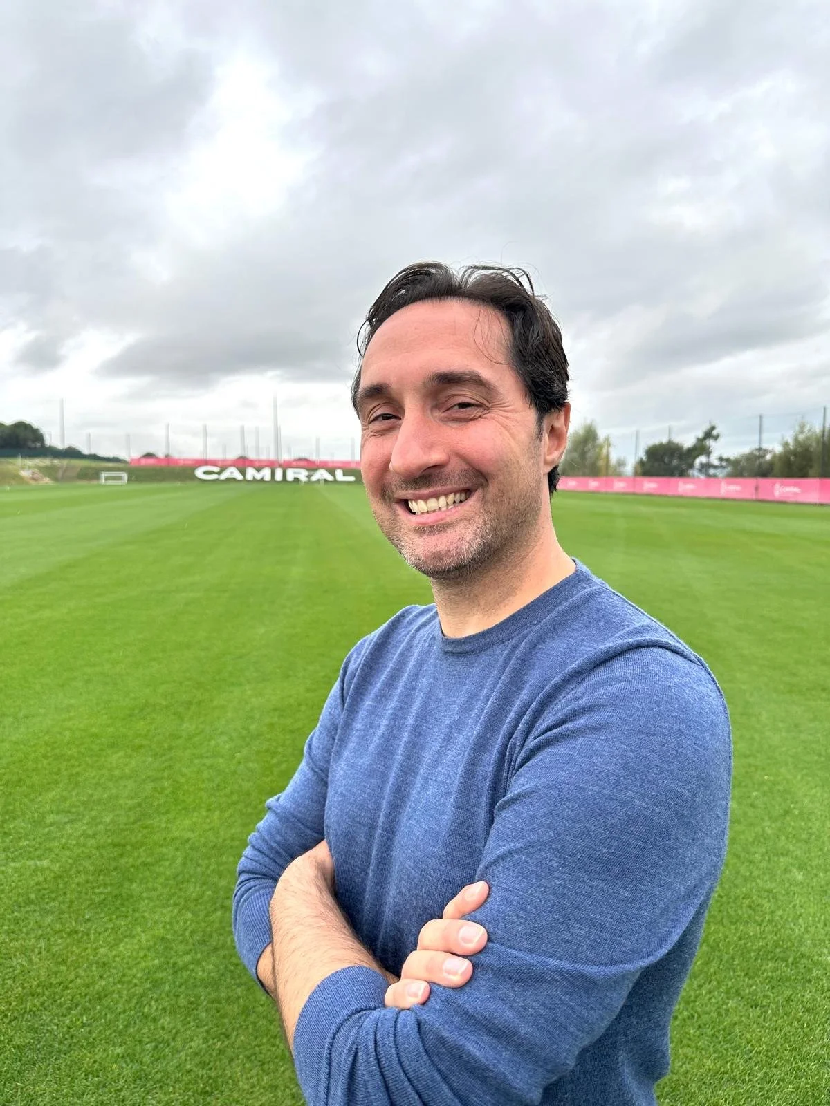 A man smiling with arms crossed on a soccer field under a cloudy sky.