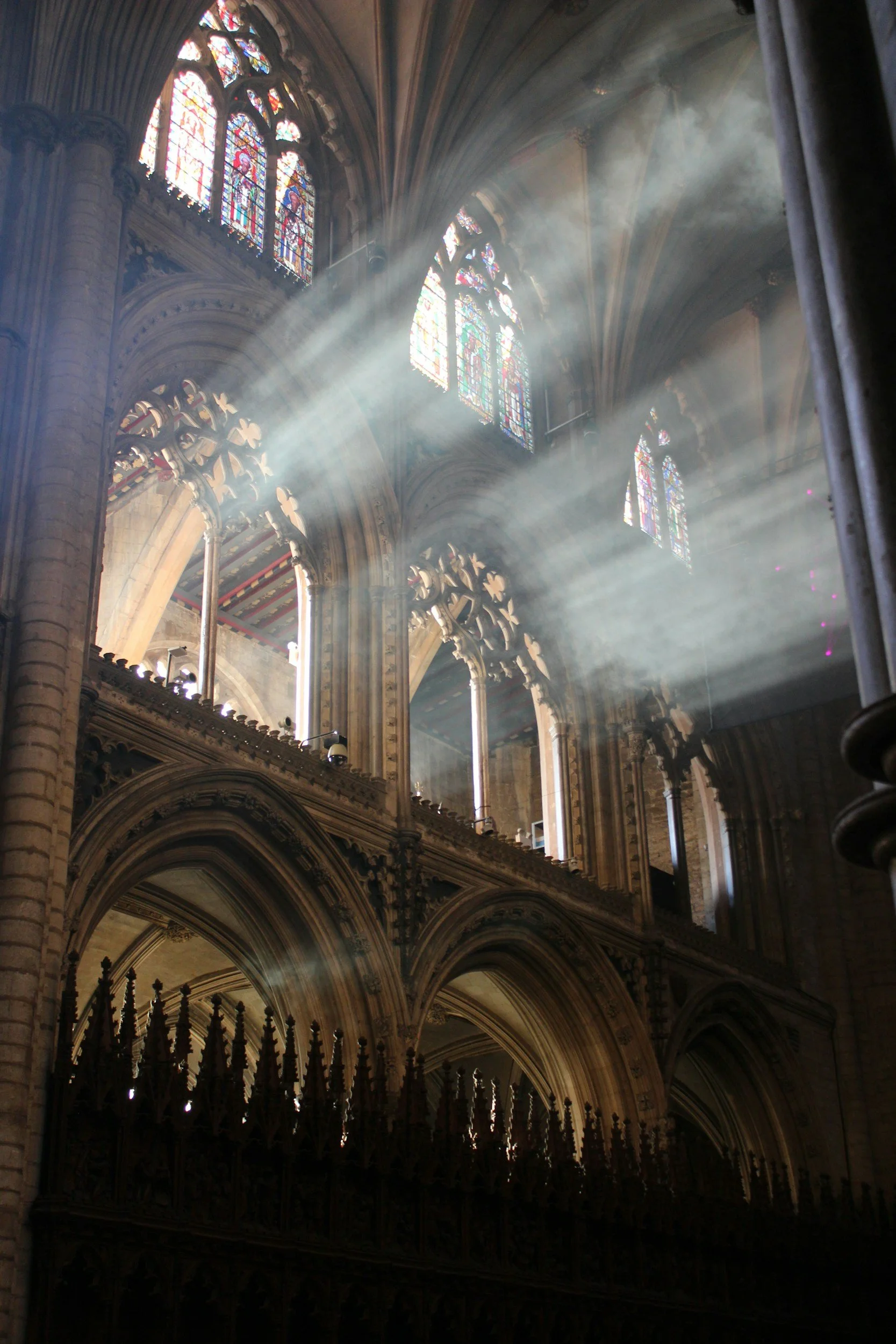 Sunlight streaming through stained glass windows inside a gothic cathedral with high vaulted ceilings and stone arches.