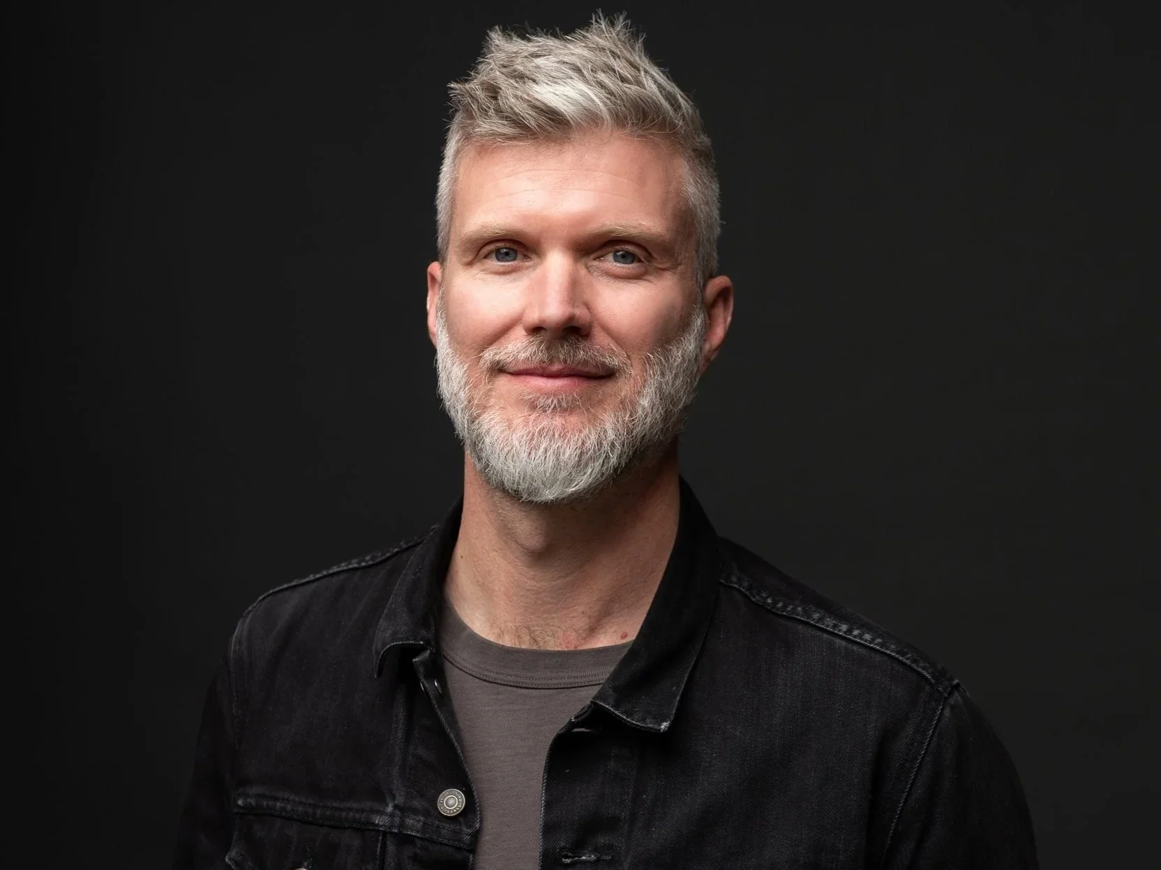 Portrait of a middle-aged man with gray hair, a beard, blue eyes, wearing a black denim jacket, against a dark background.