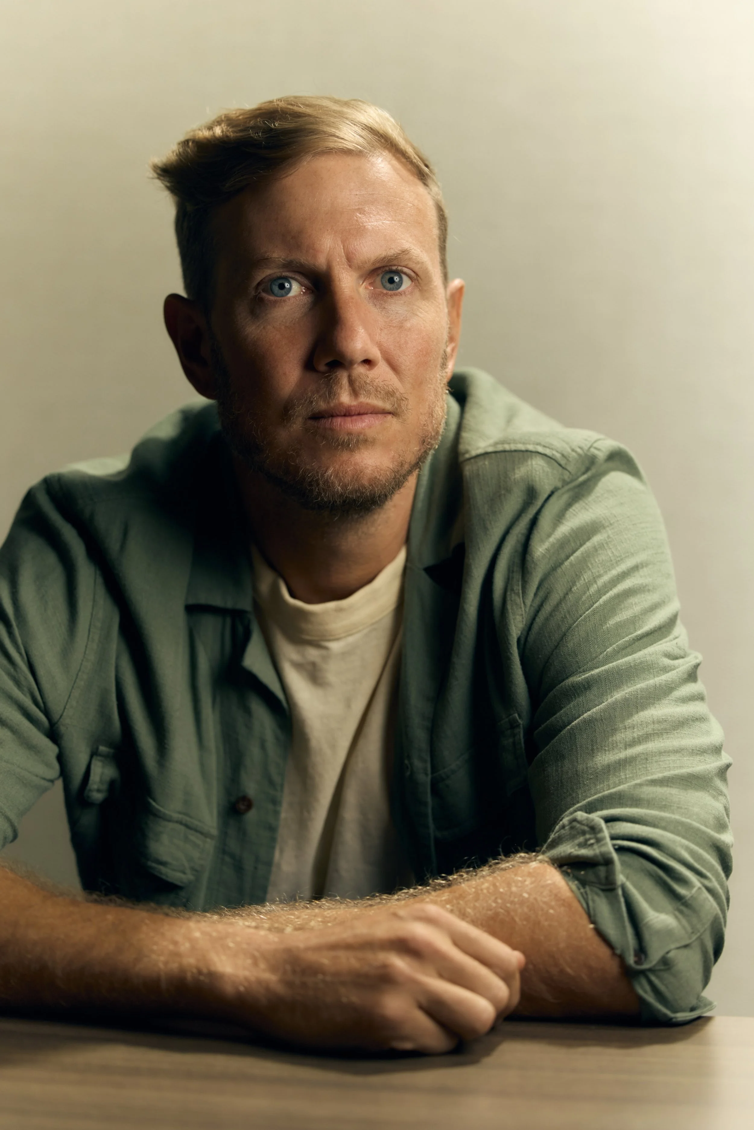 A man with short blonde hair, blue eyes, and a beard sitting at a table, wearing a green shirt over a white t-shirt, looking intently.