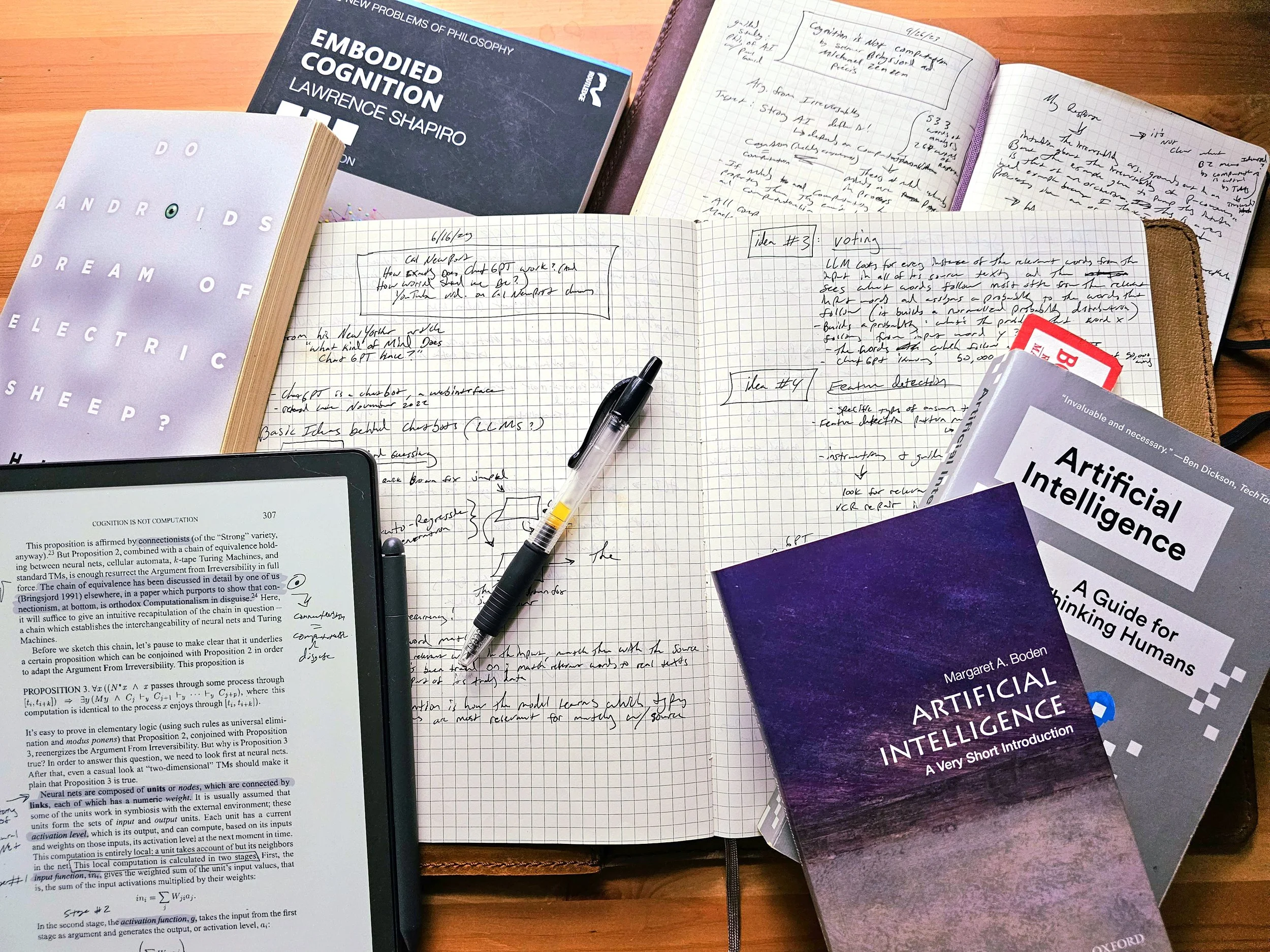 A study workspace with multiple books, notebooks, a tablet displaying a page, and handwritten notes and a pen on grid paper.