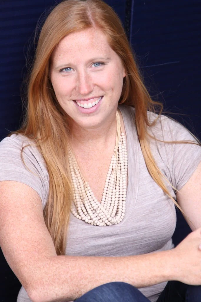 A woman with long red hair and blue eyes smiling at the camera, wearing a light gray T-shirt and a multi-strand necklace with white beads.