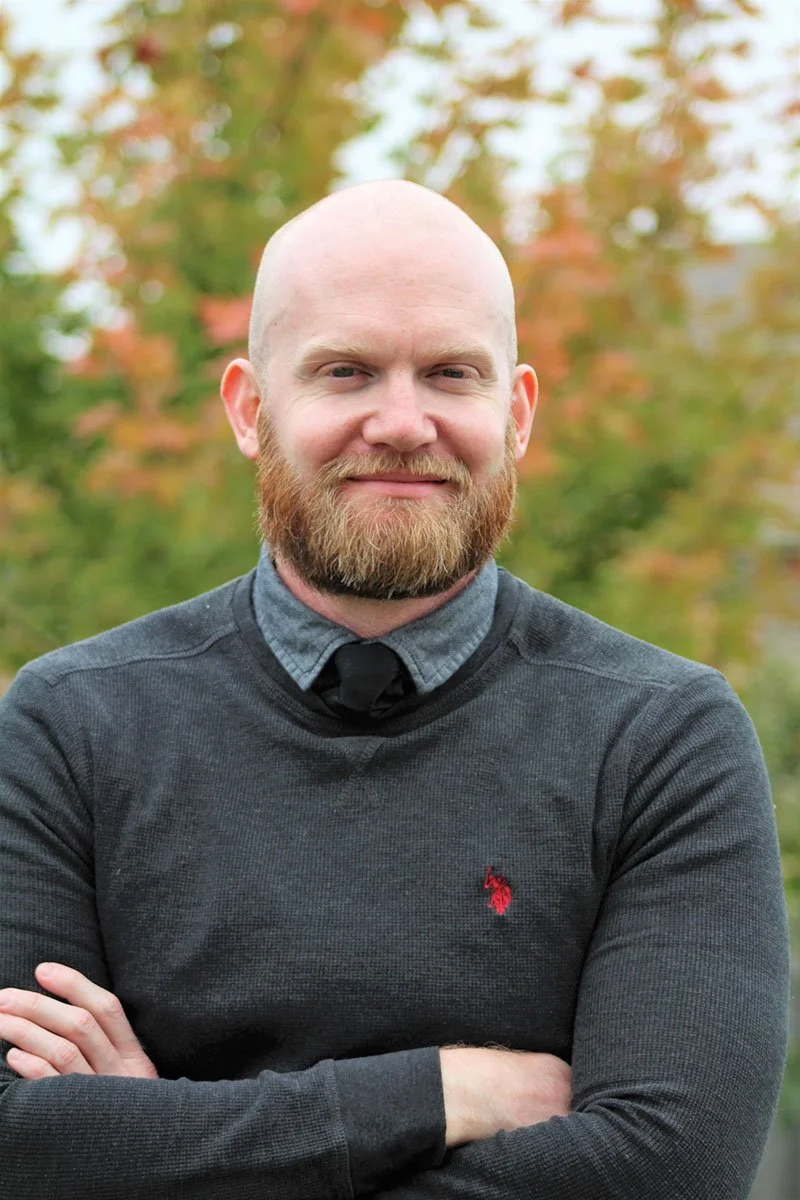 A bald man with a beard smiling outdoors, wearing a dark long-sleeve shirt and a gray collared shirt underneath, with trees in fall colors in the background.