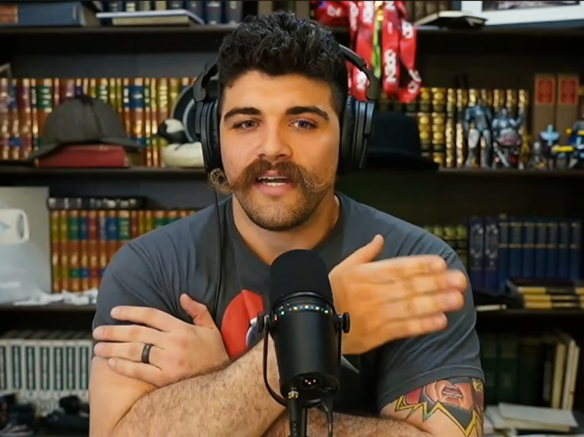 A man with a beard and mustache, wearing headphones, speaking into a microphone, gesturing with crossed arms, sitting in front of a bookshelf with books, toys, and a hat.