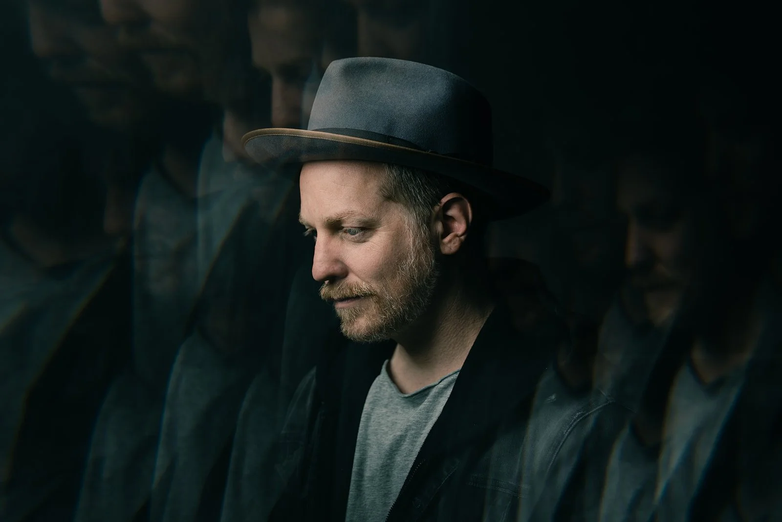 A man with a beard and light-colored hair wearing a gray hat, gray T-shirt, and black jacket, looking down with a reflective expression, surrounded by blurry reflections of himself.