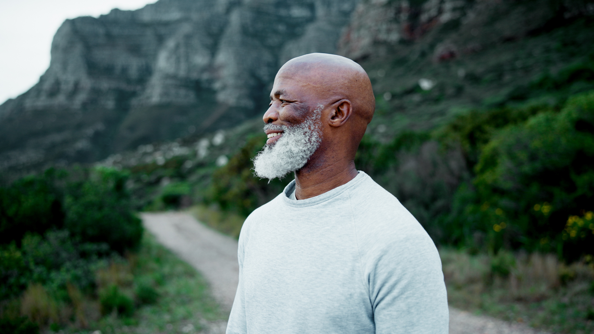An elderly man with a white beard and a bald head standing outdoors on a dirt trail, with greenery and rocky hills in the background.