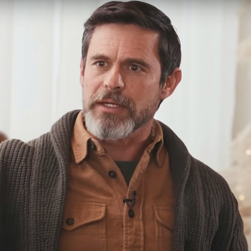 A middle-aged man with dark hair and a gray beard speaking, wearing a brown button-up shirt and a dark cardigan, in an indoor setting.