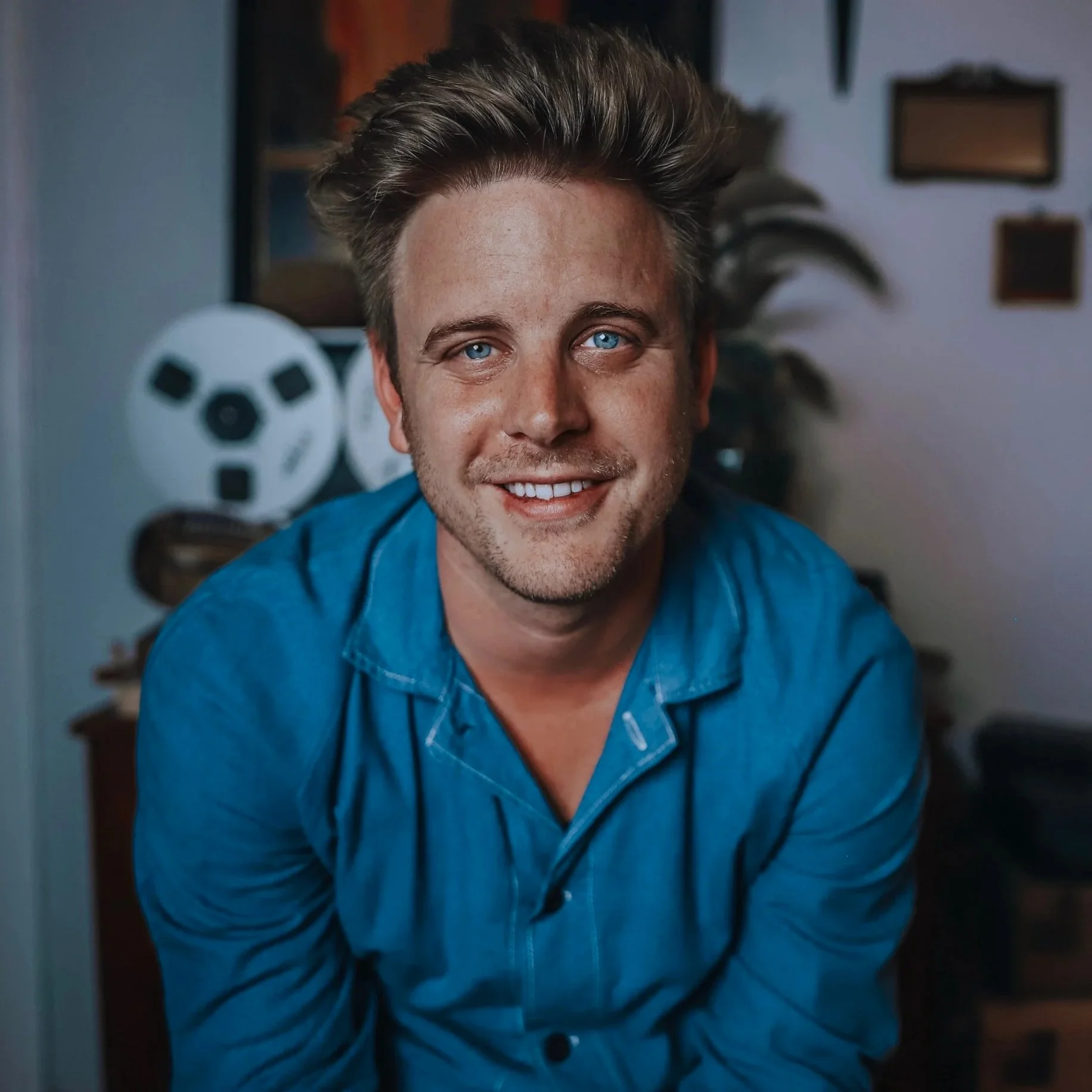 A smiling young man with blue eyes and wavy brown hair, wearing a blue chambray shirt, sitting indoors with a film reel, a large plant, and picture frames in the background.