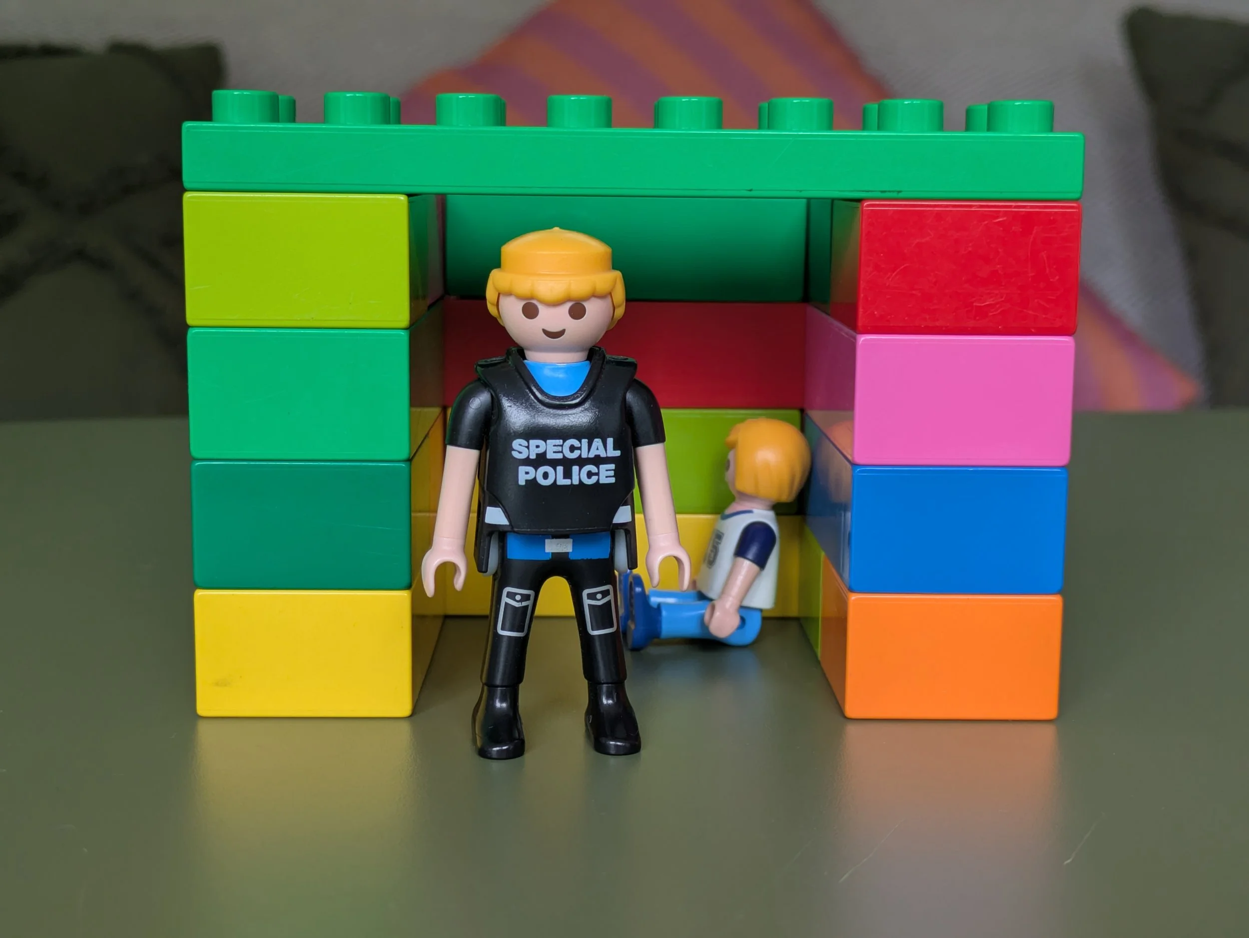 Duplo bricks made into a building with a playmobile child sat inside and a policeman guarding the entrance