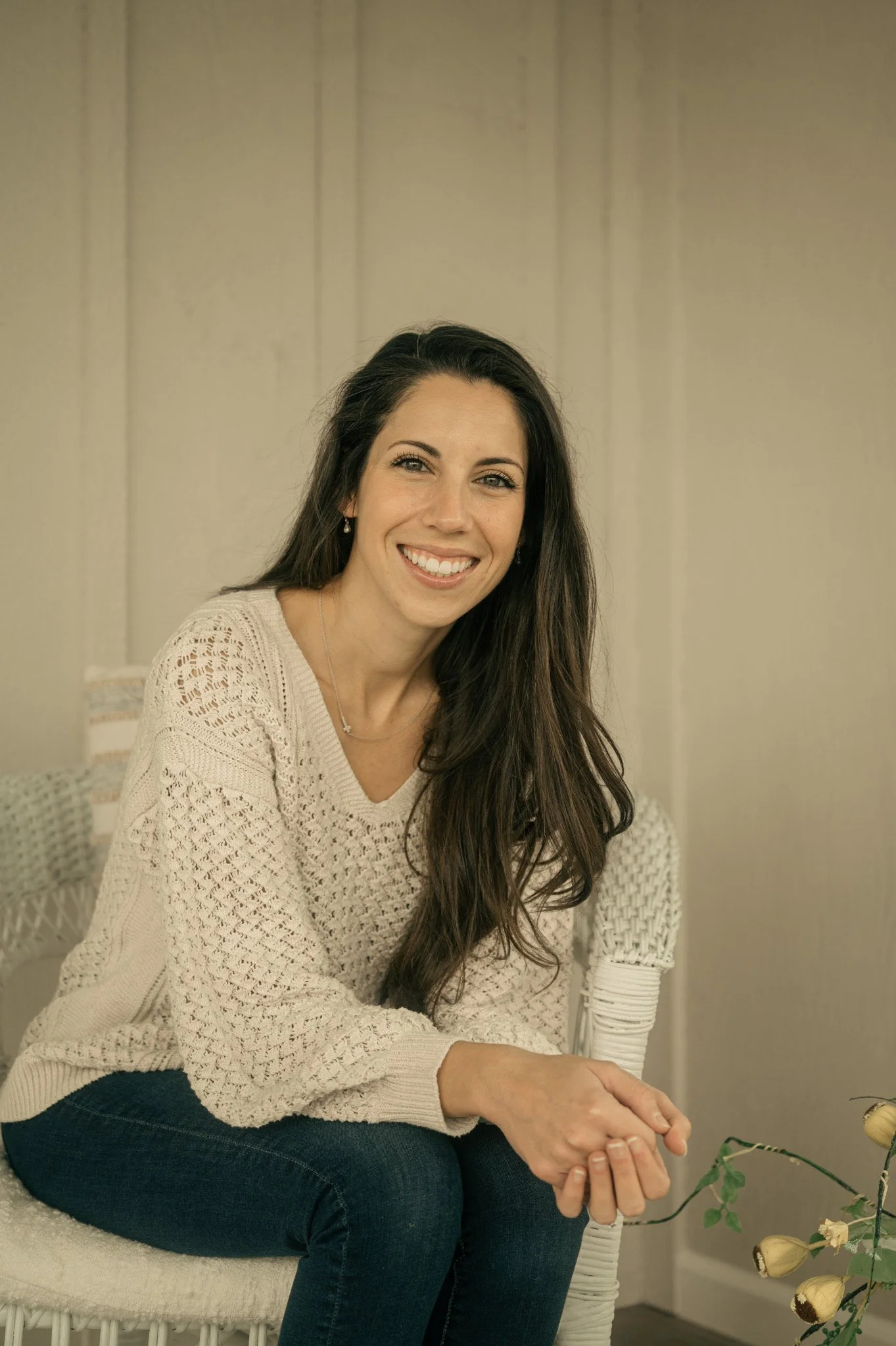 A woman with long dark hair and a light sweater sitting on a white wicker chair, smiling at the camera in a neutral-colored room.