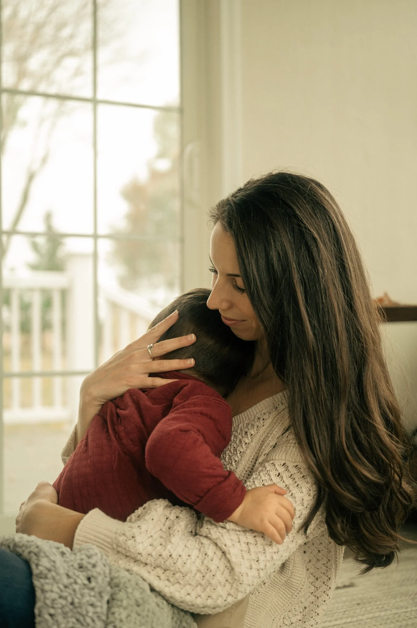 A woman hugging and comforting a young child inside a home near a window.
