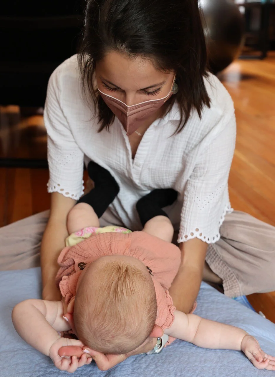 A woman in a white blouse and face mask holding a sleeping baby upside down on a bed.