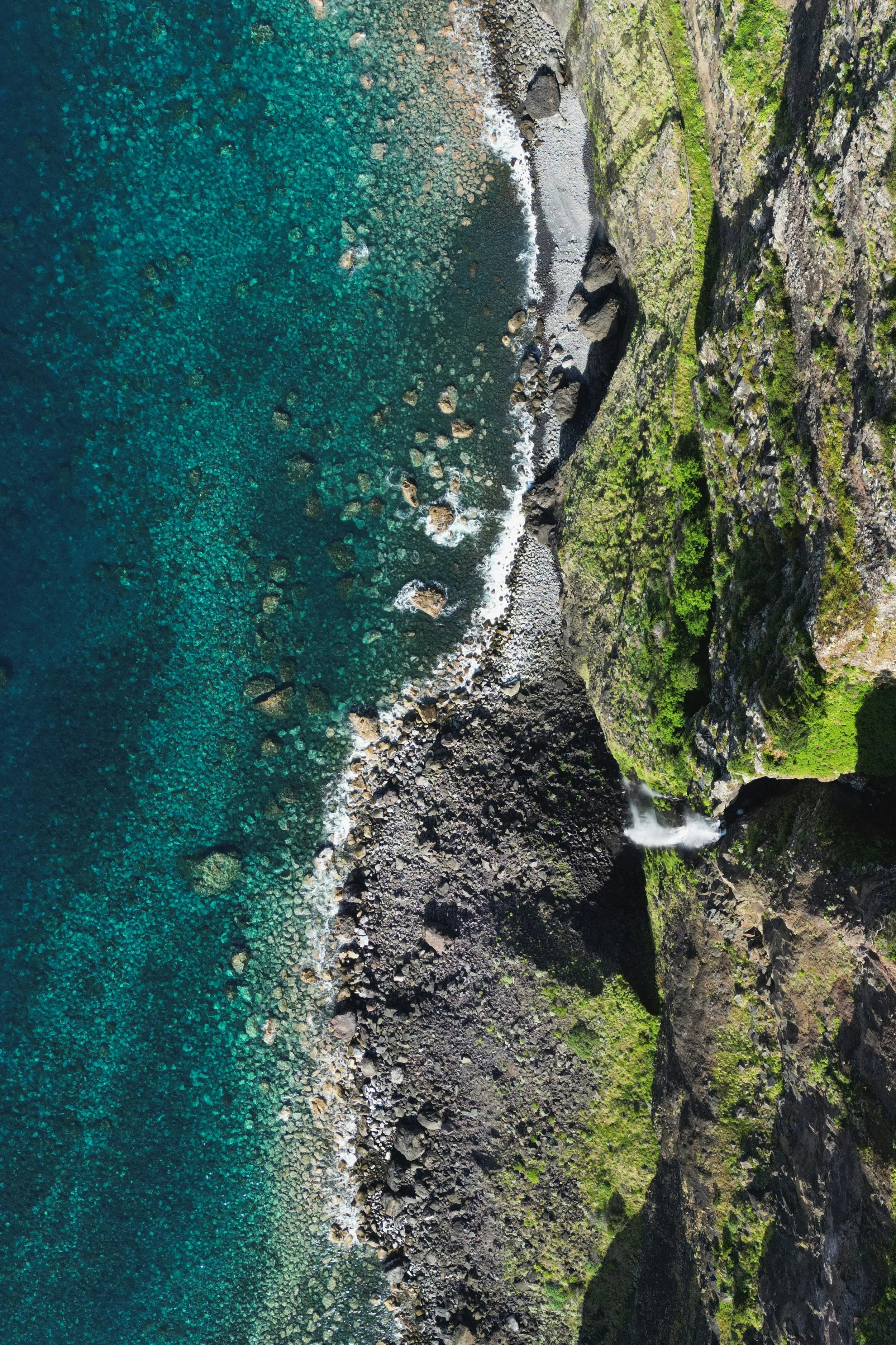 Aerial view of a Hawaiian rugged coastline with a steep green hillside, rocky shore, and turquoise ocean with scattered rocks and waves.