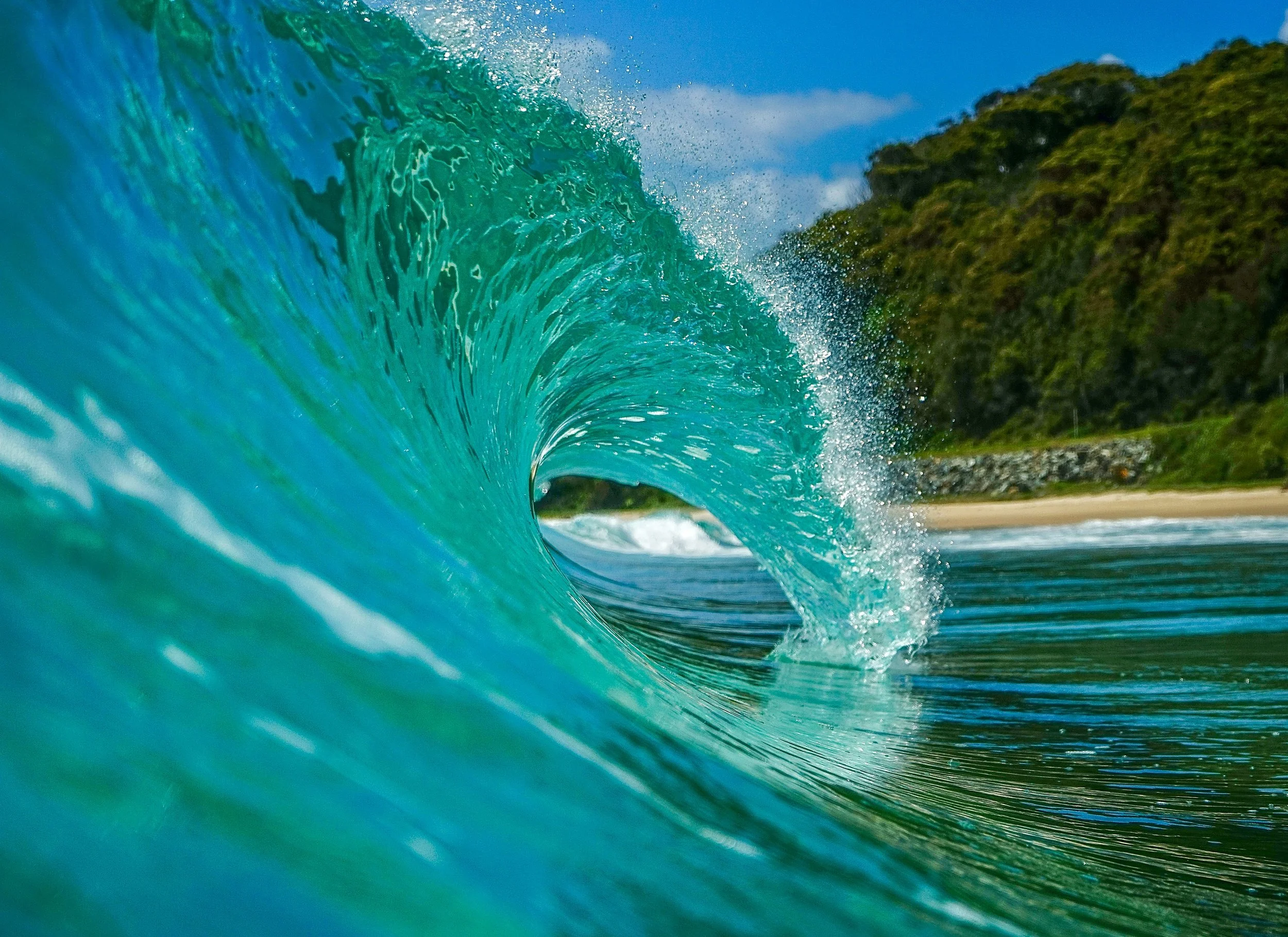A vibrant ocean wave curling near a Hawaiian shoreline, green forested hill in the background.