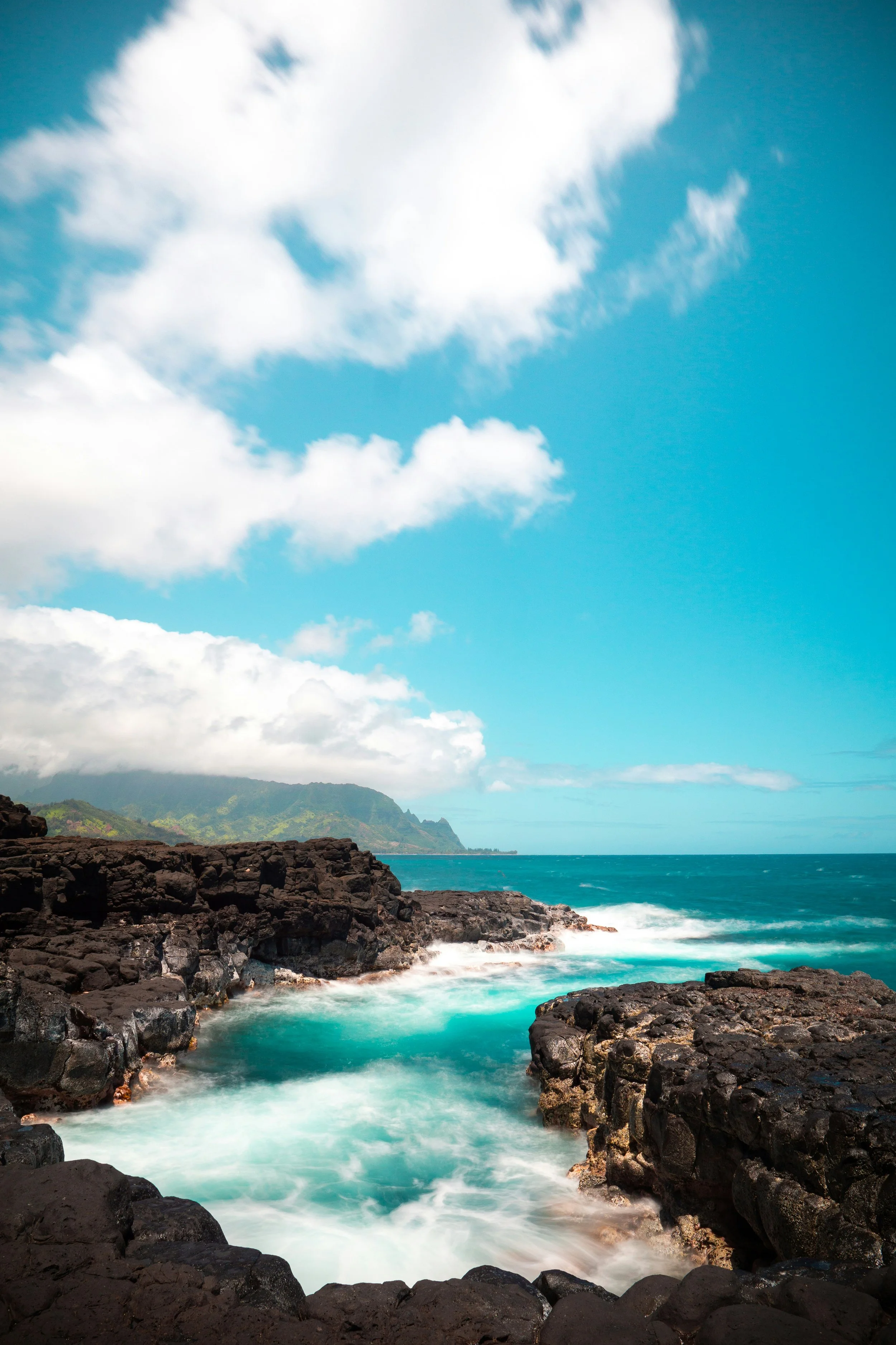 Hawaiian tropical coastline with black volcanic rocks, turquoise water, and a partly cloudy sky over distant green mountains.