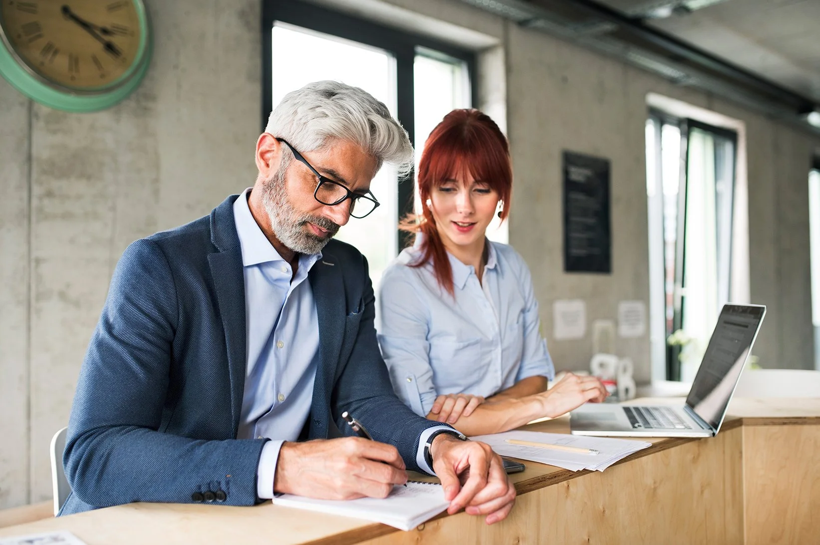 Two professionals, a man with gray hair and a woman with red hair, sitting at a desk in an industrial-style office, working with documents and a laptop.