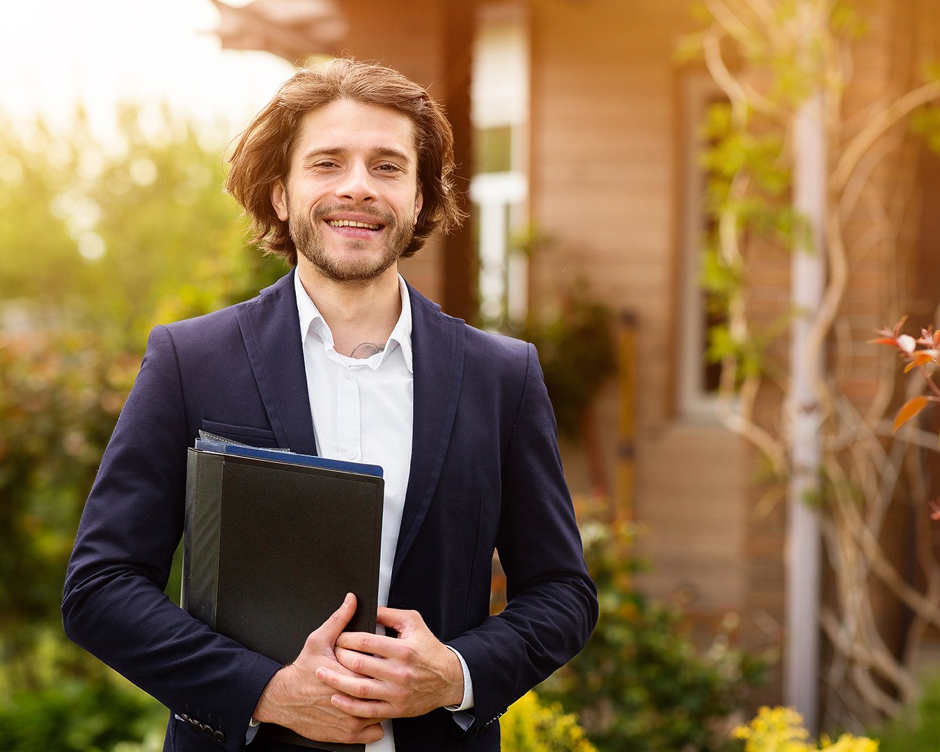 A man smiling outdoors, dressed in a dark blazer and white shirt, holding folders, with a background of greenery and a building.