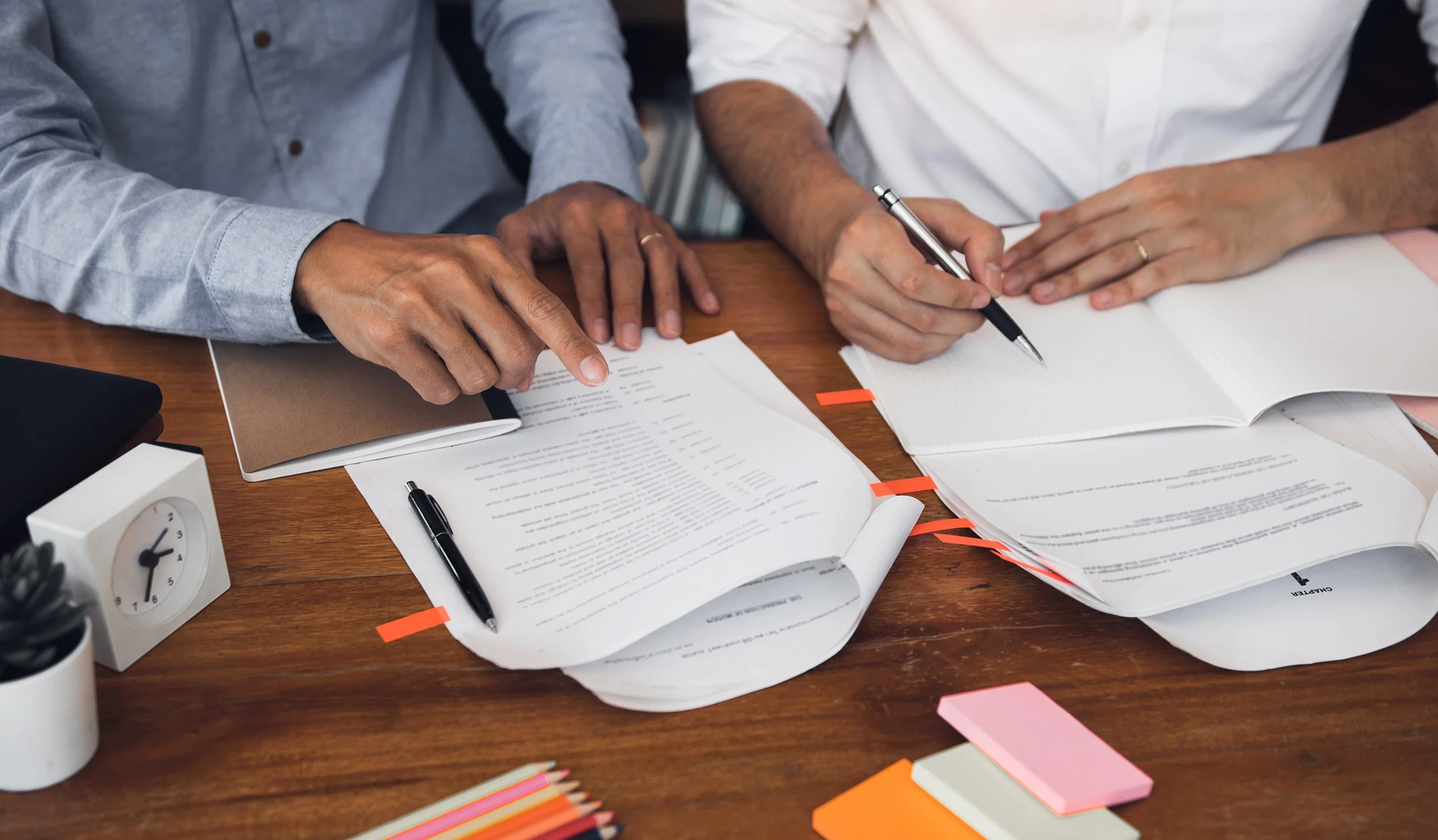 Two individuals discussing documents at a wooden table, with notebooks, pens, sticky notes, and a small clock on the table.
